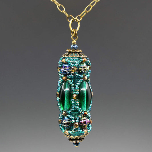 A turquoise and green beaded pendant hangs from a gold colored chain. The pendant has long oval beads that form the center row and textured iridescent beads that form a decorative top and bottom row. A netting of translucent peacock green seed beads is overlaid, outlining all of the larger beads.