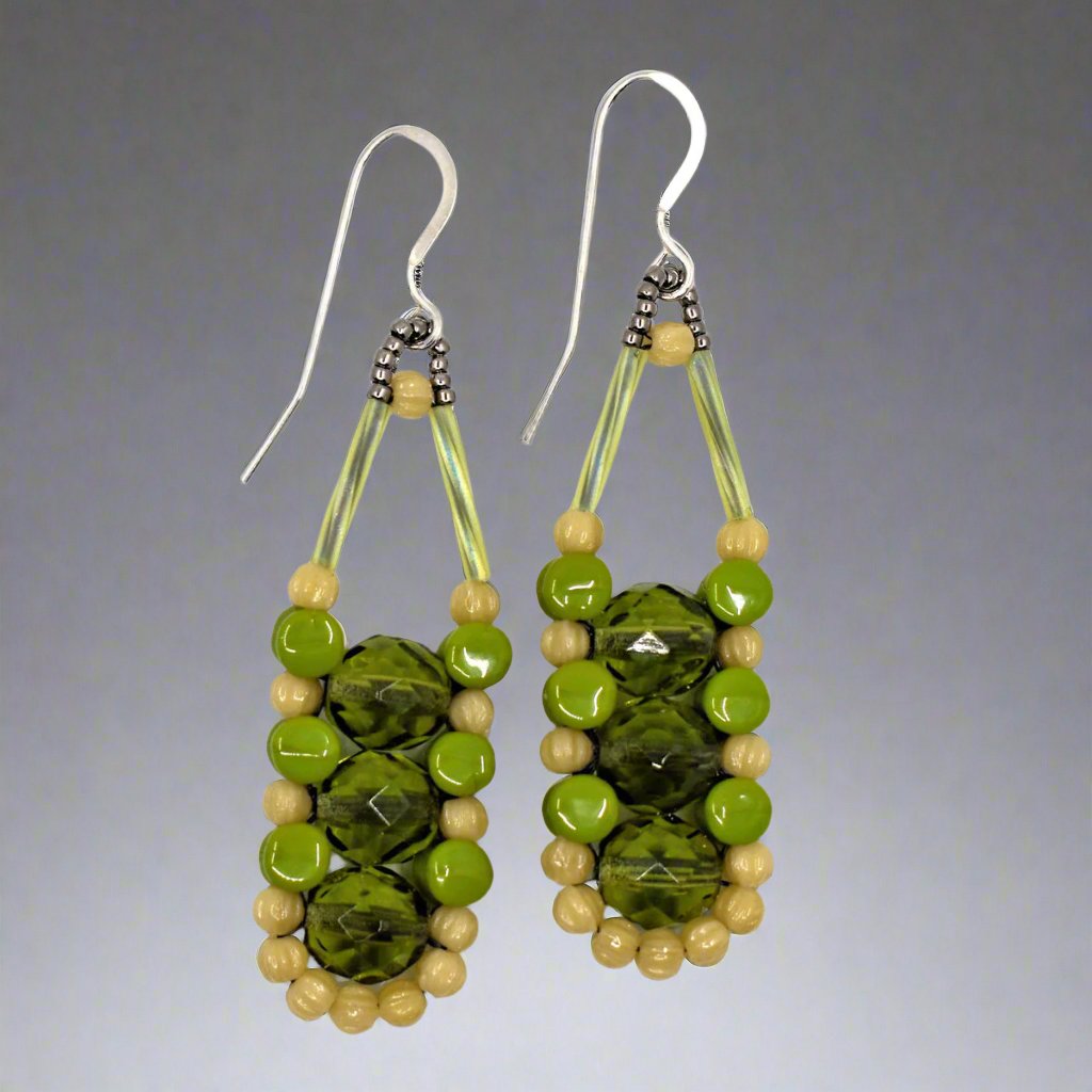 Long green earrings with dark cream and avocado, with silver accents and ear wires on a gray background. There are three olive green beads stacked vertically in the center of these beads, like peas, surrounded by an outline of dark cream and avocado green beads.