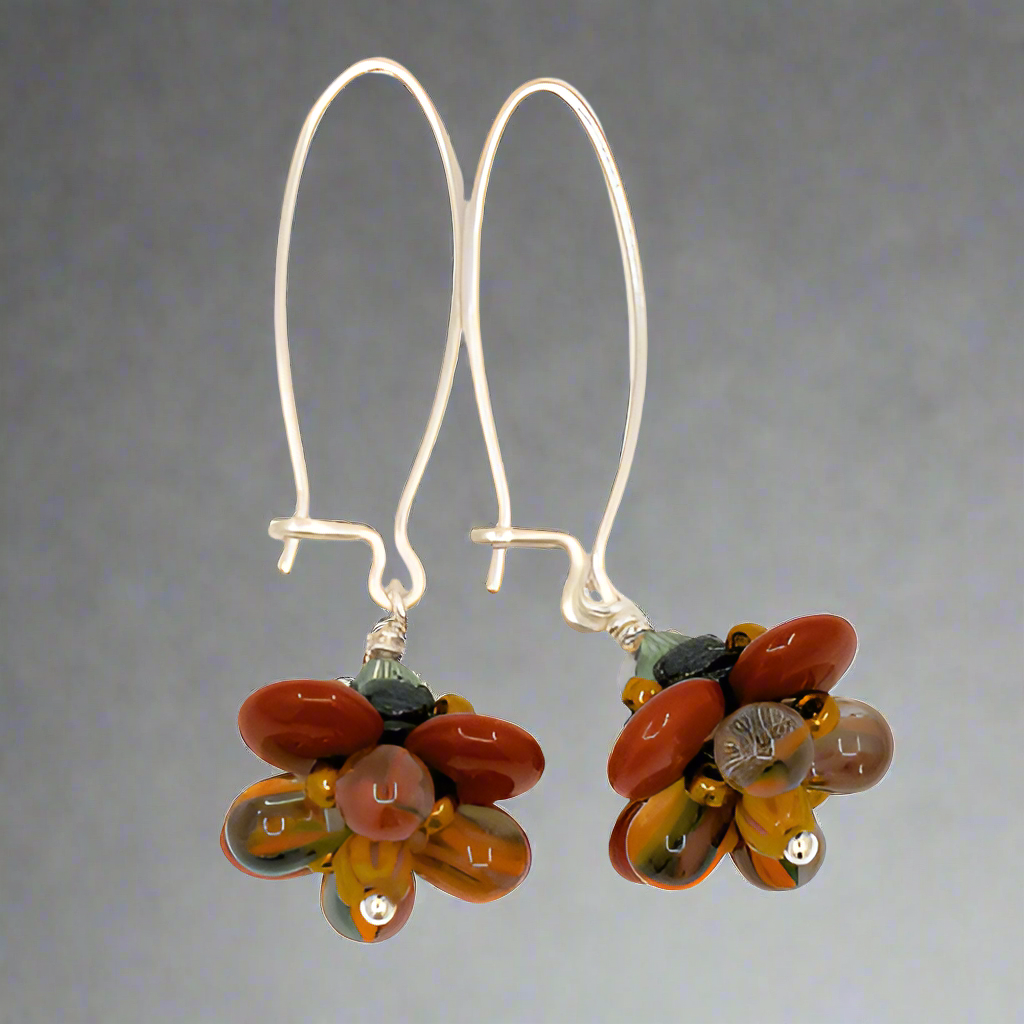 A pair of silver earrings with a brown, mustard and green flower on the bottom against a gray background. The earrings have long silver oval wires that latch and the dangling parts are beaded flowers with rust colored outer petals and interior petals that are clear with mustard, dark green, and brown stripes.
