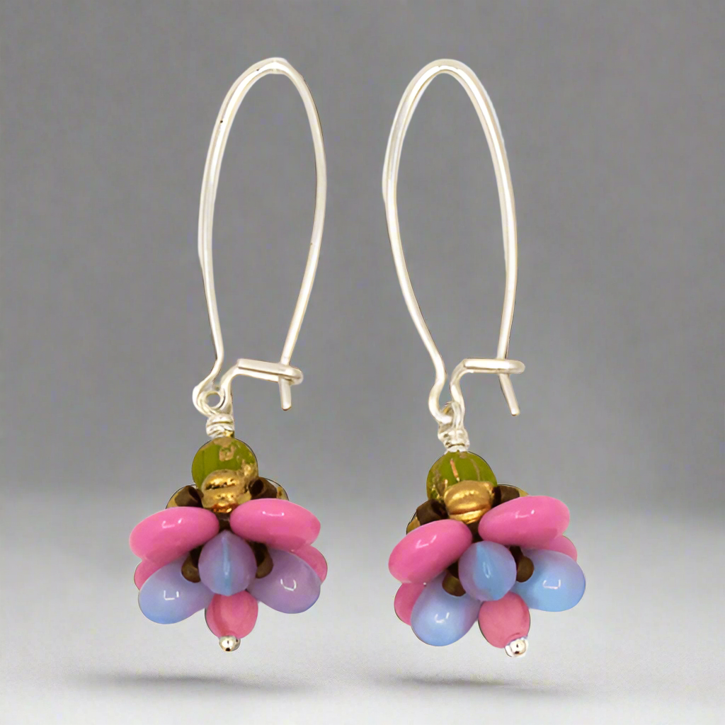A pair of silver earrings with pastel pink and purple flower drops against a gray background. The earrings have long silver oval wires that latch and the dangling parts are beaded flowers with pink outer petals and pale purplish blue inner petals.
