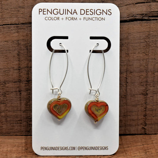 A pair of earrings on a white card rest against a wood background. The earrings have long silver oval wires that latch with heart beads at the bottom that are a vaiegated dark orange and yellow. The hearts are cut to show the colored glass in a wide outline around the outer part, leaving the outside edges and embossed interior heart shape covered in the stone-like tan finish