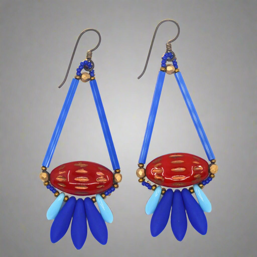 Long blue and red earrings shaped like a teardrop with tail feathers lay on a gray background. The upper part of the teardrop shape is formed by long medium blue tubes. At the base is a glossy red pointed oval with copper horizontal dashes. At the bottom is a spray of dagger shaped beads, the center three are matte medium blue and the outer two are shiny and light blue. The earrings have dark ear wires.