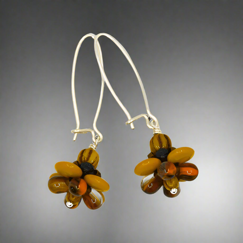 A pair of silver earrings with mustard and brown flowers against a gray background. The earrings have long silver oval wires that latch and dangling beaded flowers with mustard outer petals and inner petals made of clear glass with orange and brown stripes inside of it..