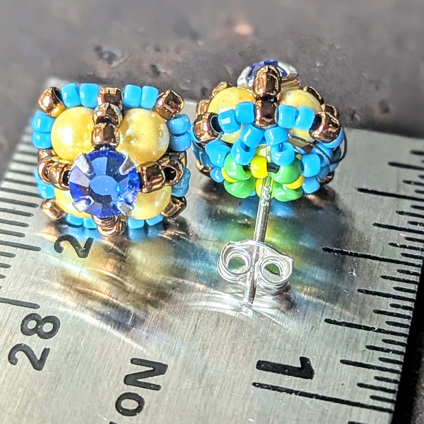 A pair of four-lobed stud earrings with yellow beads surrounded by smaller turquoise beads and a blue rhinestone center, on a ruler for scale. One earring is shown from the back, which has green bead detailing.