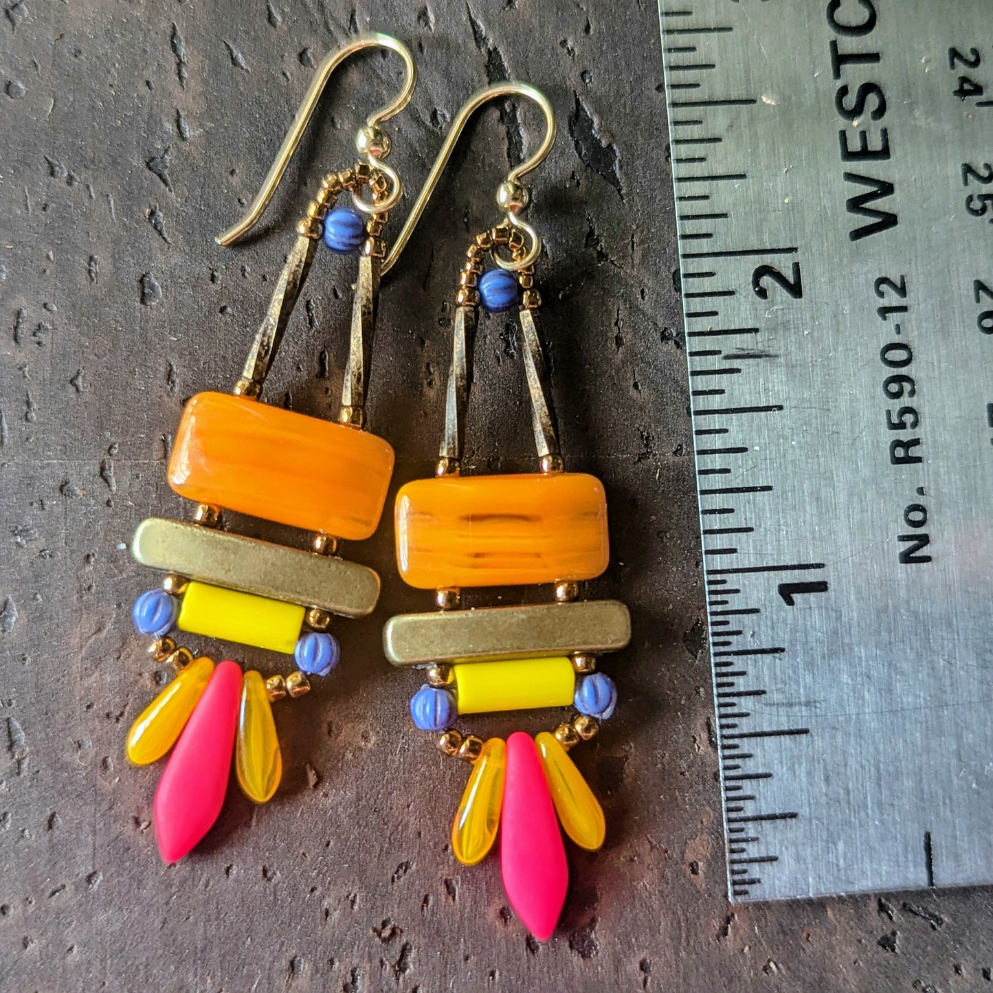 Brightly colored earrings with orange, pink, yellow and blue beads, shown with a ruler for scale on a dark textured surface