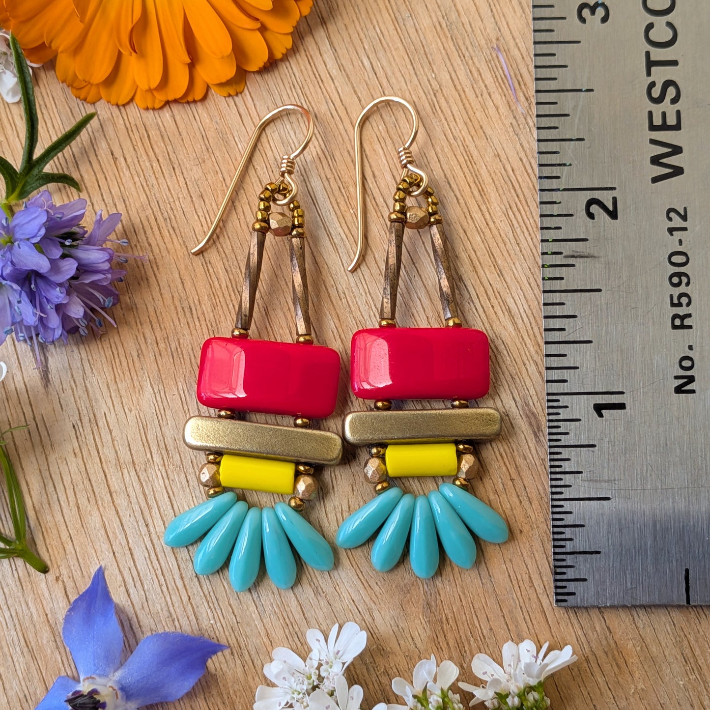 colorful earrings on a wood background with flowers and a ruler for scale. The earrings each have a red rectangle stacked on top of a brass-colored bar. Under the bar there is light yellow tube, five small turquoise daggers and tiny gold accents.