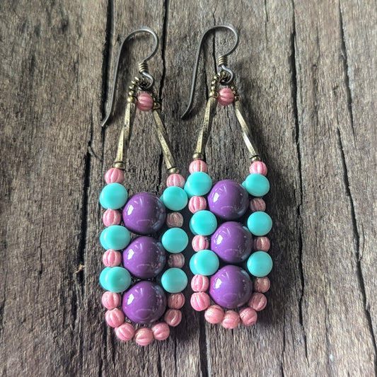 Columnar beaded earrings with purple, turquoise, and pink beads on a wooden surface.