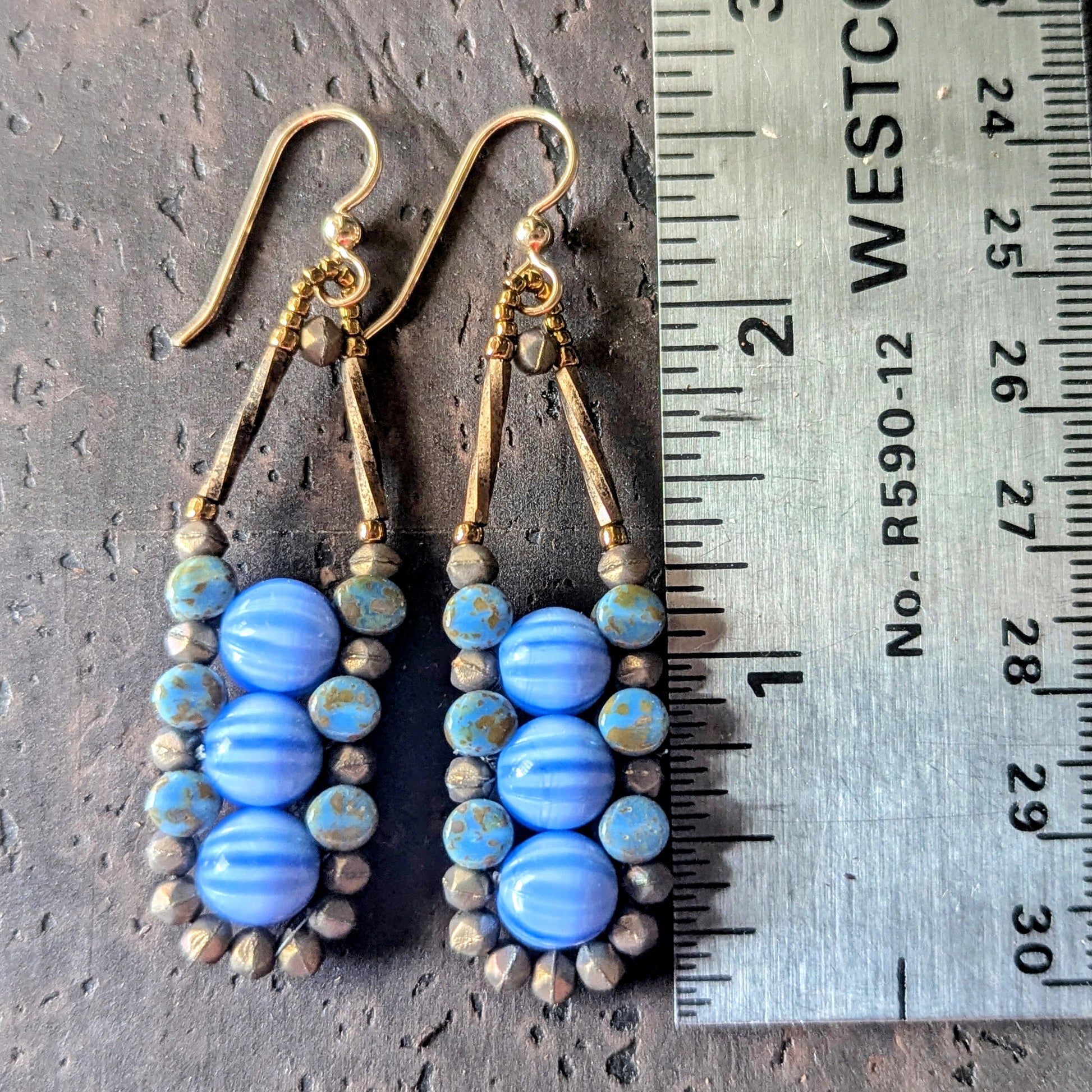 Pair of blue beaded earrings with gold accents on a dark surface next to a ruler.