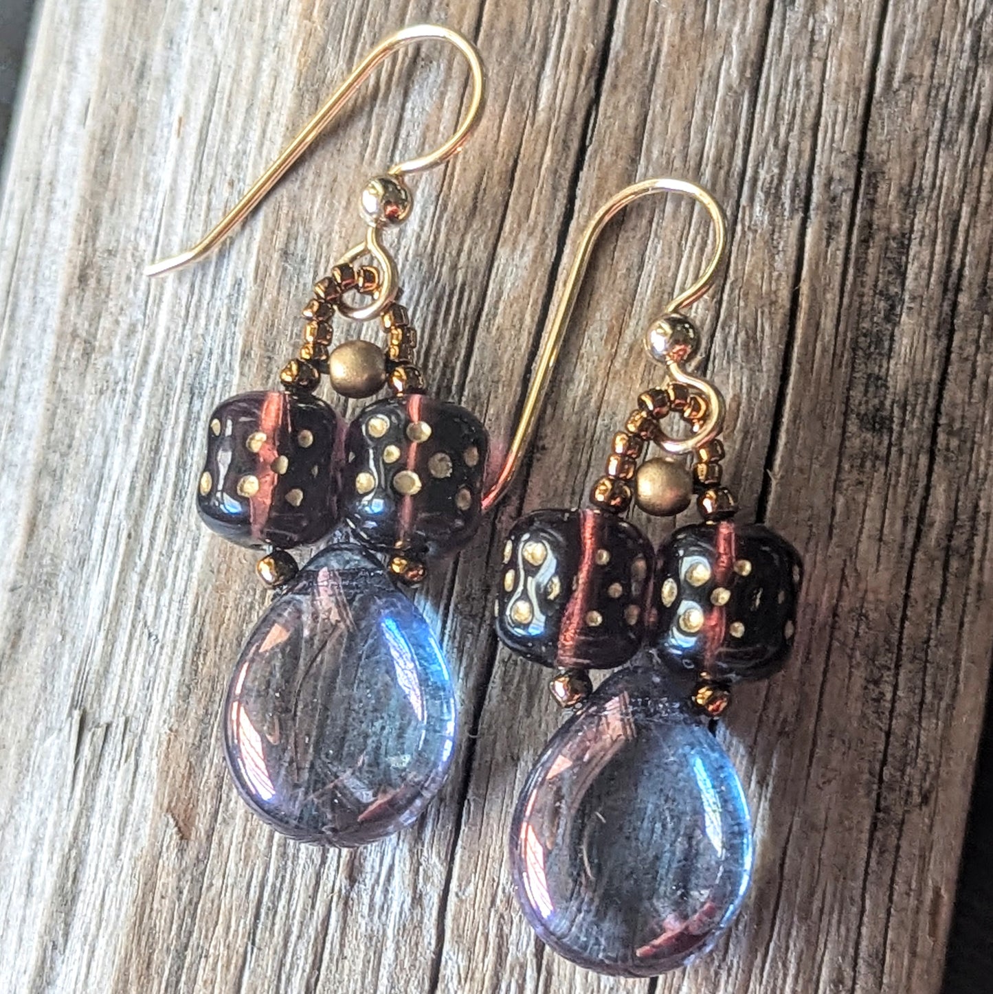 Pair of earrings with purple teardrop beads, plum cylinder beads, and gold accents on a wooden surface.