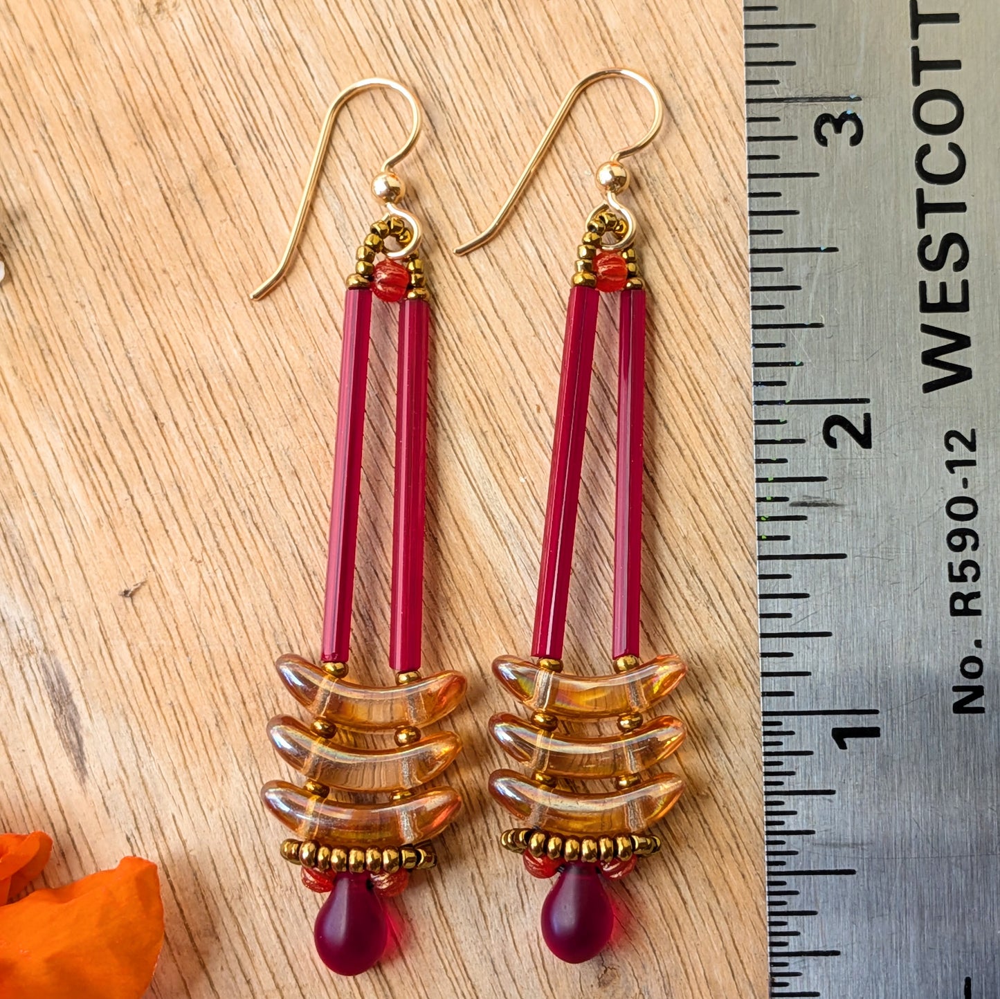 Long red an apricot earrings on a wood background with orange flowers and a ruler for scale.