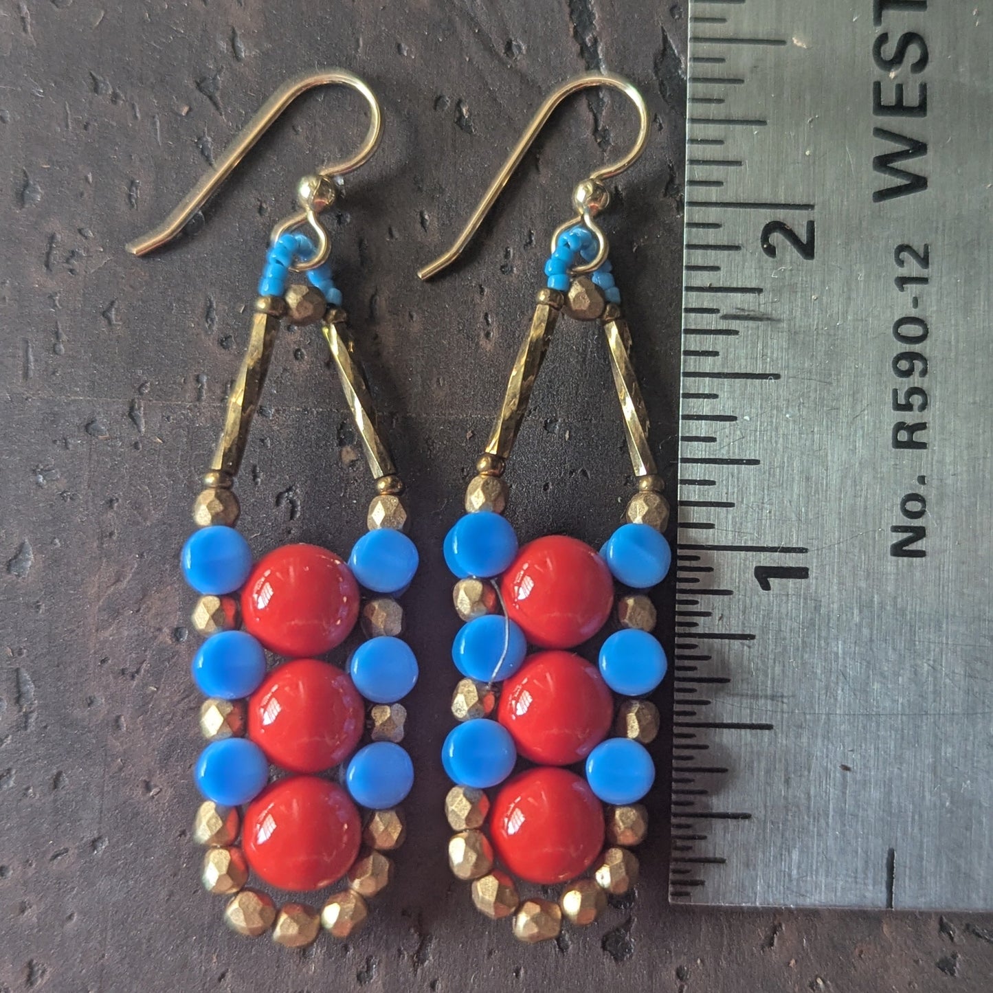 A columnar pair of beaded earrings with red, blue and gold beads on a wooden surface, shown next to a ruler for scale.
