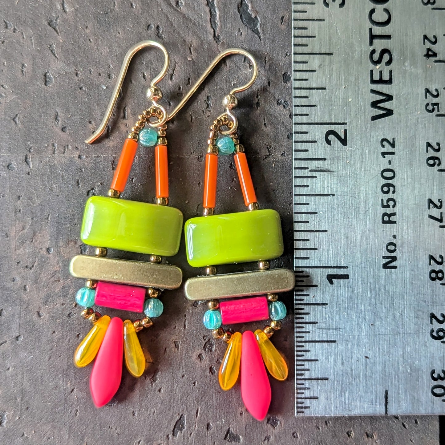 Brightly colored earrings with green, pink. orange ant teal on a textured background with a ruler for scale.