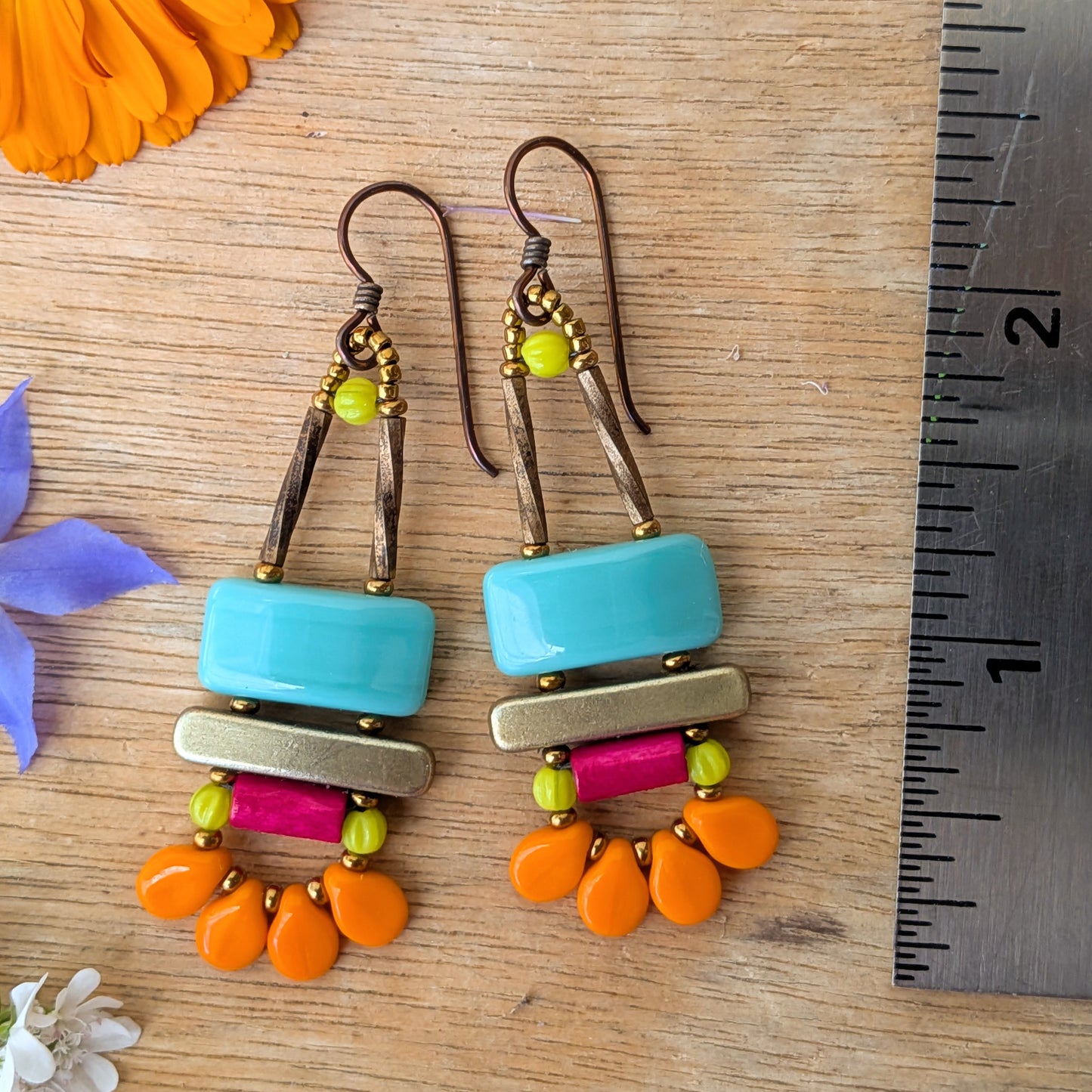 Colorful earrings on a wooden surface with a ruler for scale. The earrings have turquoise rectangles on top, a gold bar in the middle and a magenta tube with a swag of petal-like orange beads on the bottom.