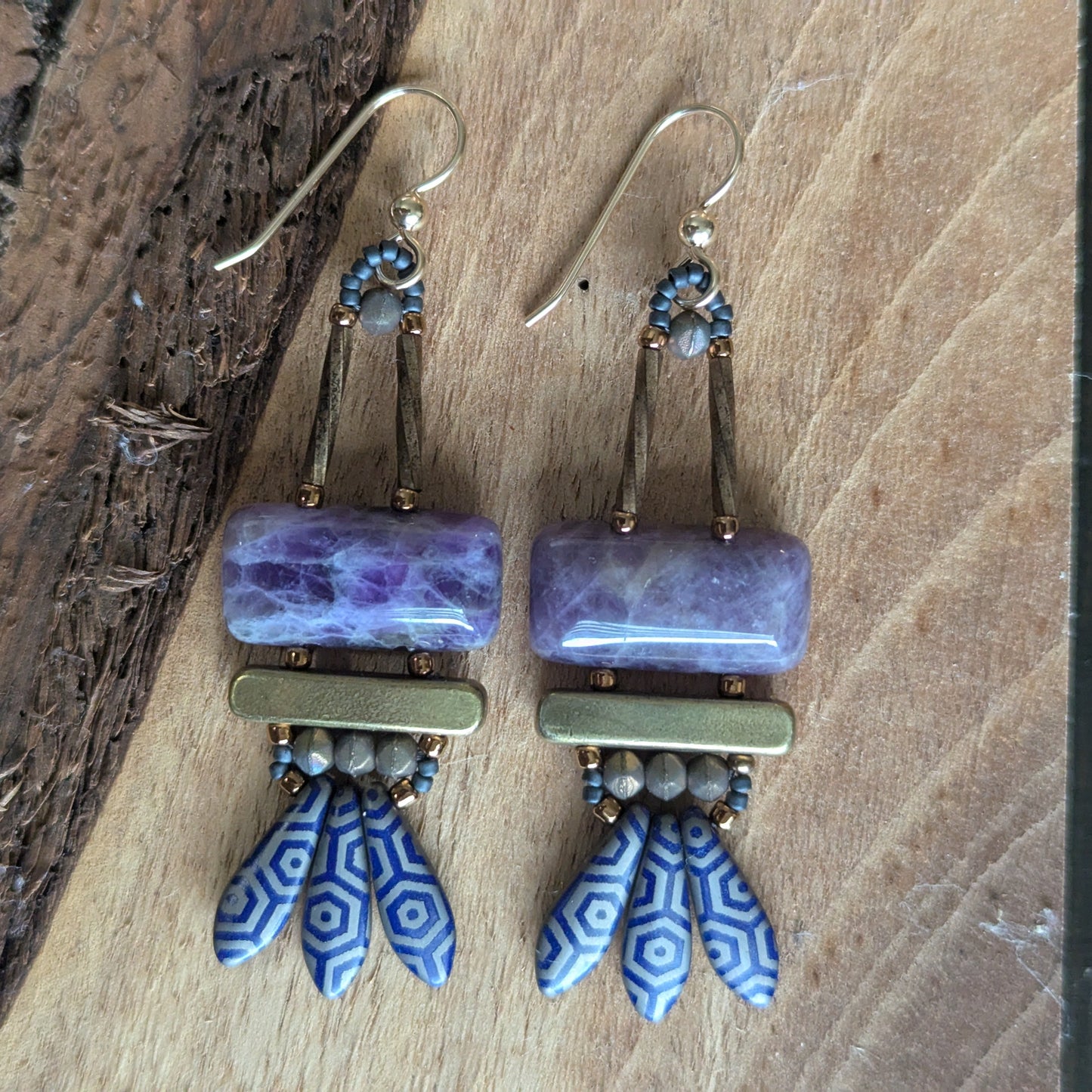 Purple earrings on a slice of wood. The earrings have purple stone rectangles on top with gold bars and navy blue daggers with a silver-gray octagon pattern.