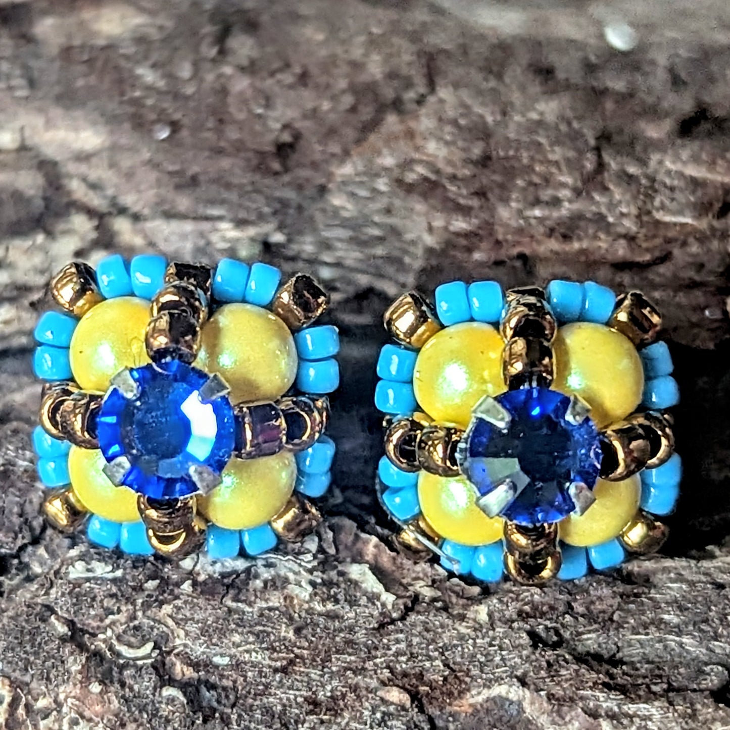 Four-lobed stud earrings with yellow beads surrounded by smaller turquoise beads and a blue rhinestone center, on a wood bark background.