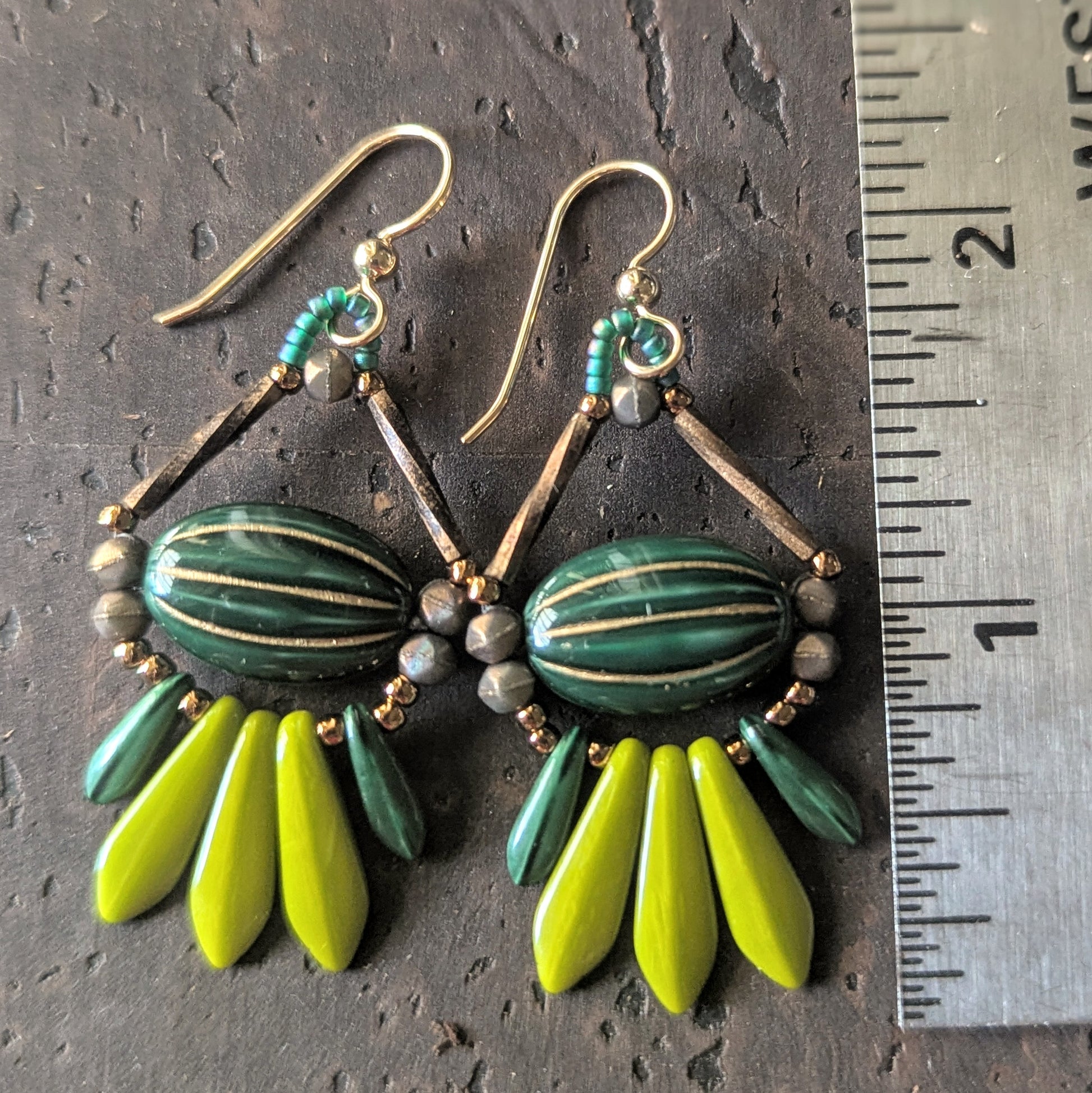 Green and gold earrings with a ruler for scale on a dark brown surface