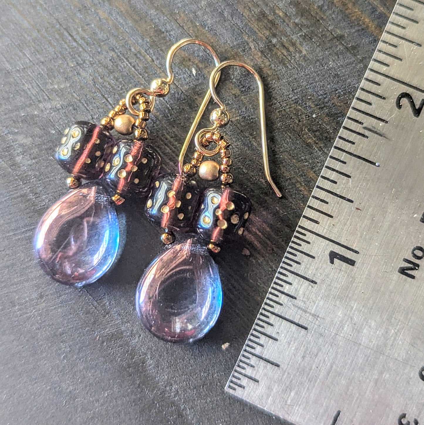 Purple teardrop earrings with gold accents on a dark surface next to a ruler.