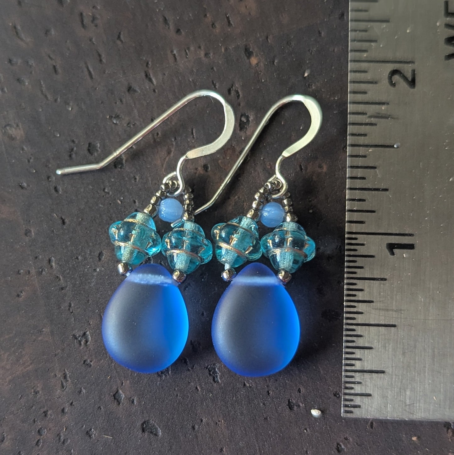 Blue teardrop earrings with aqua beads on a dark surface with a ruler for scale.