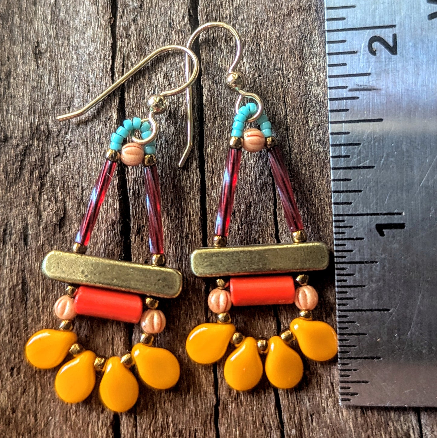 Colorful beaded earrings with shades of orange, gold, and turquoise on a wooden surface with a ruler for scale.