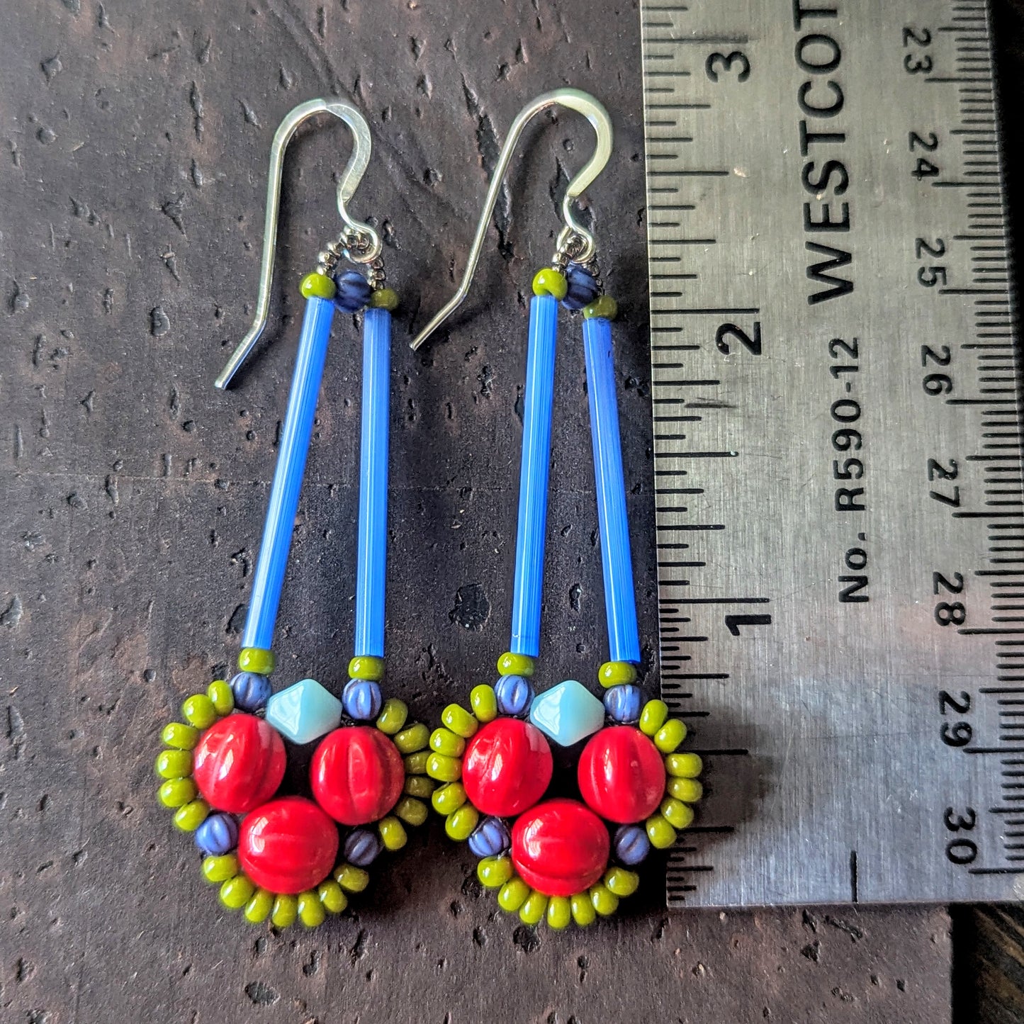 Long earrings with red, green and blue beads on a wooden surface, shown with a ruler for scale.