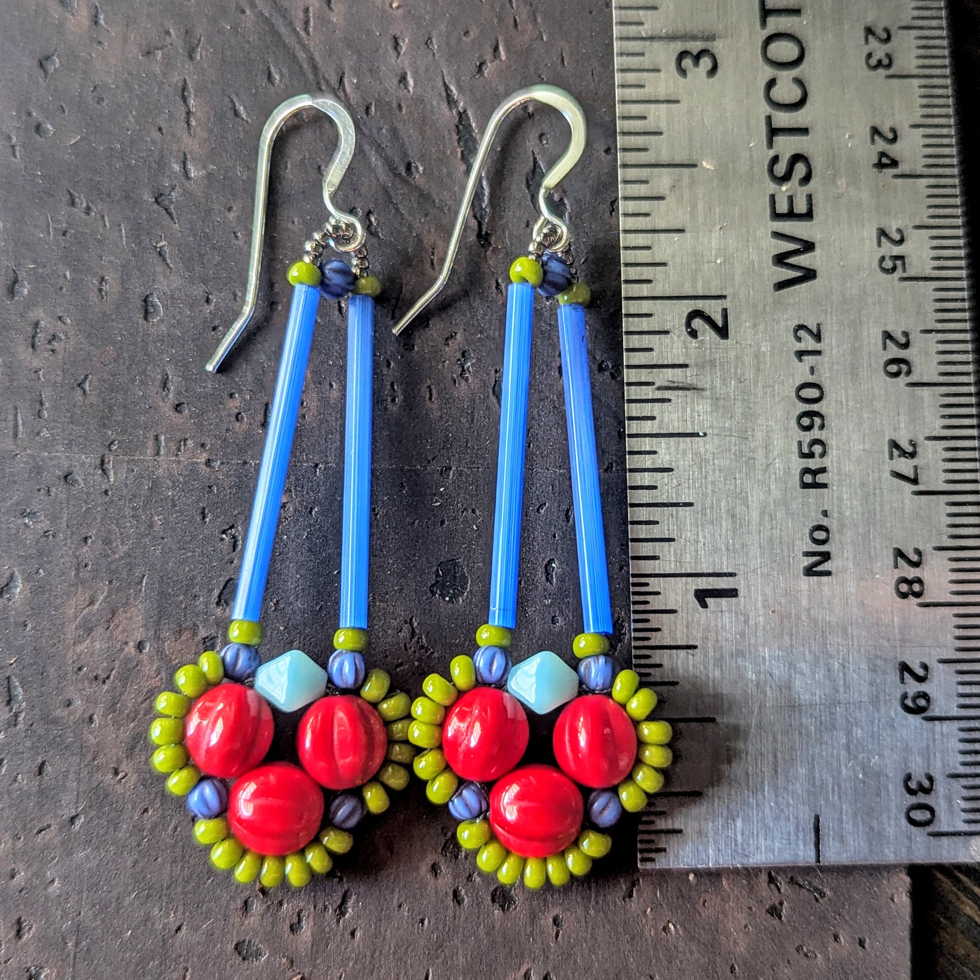 Long earrings with red, green and blue beads on a wooden surface, shown with a ruler for scale.