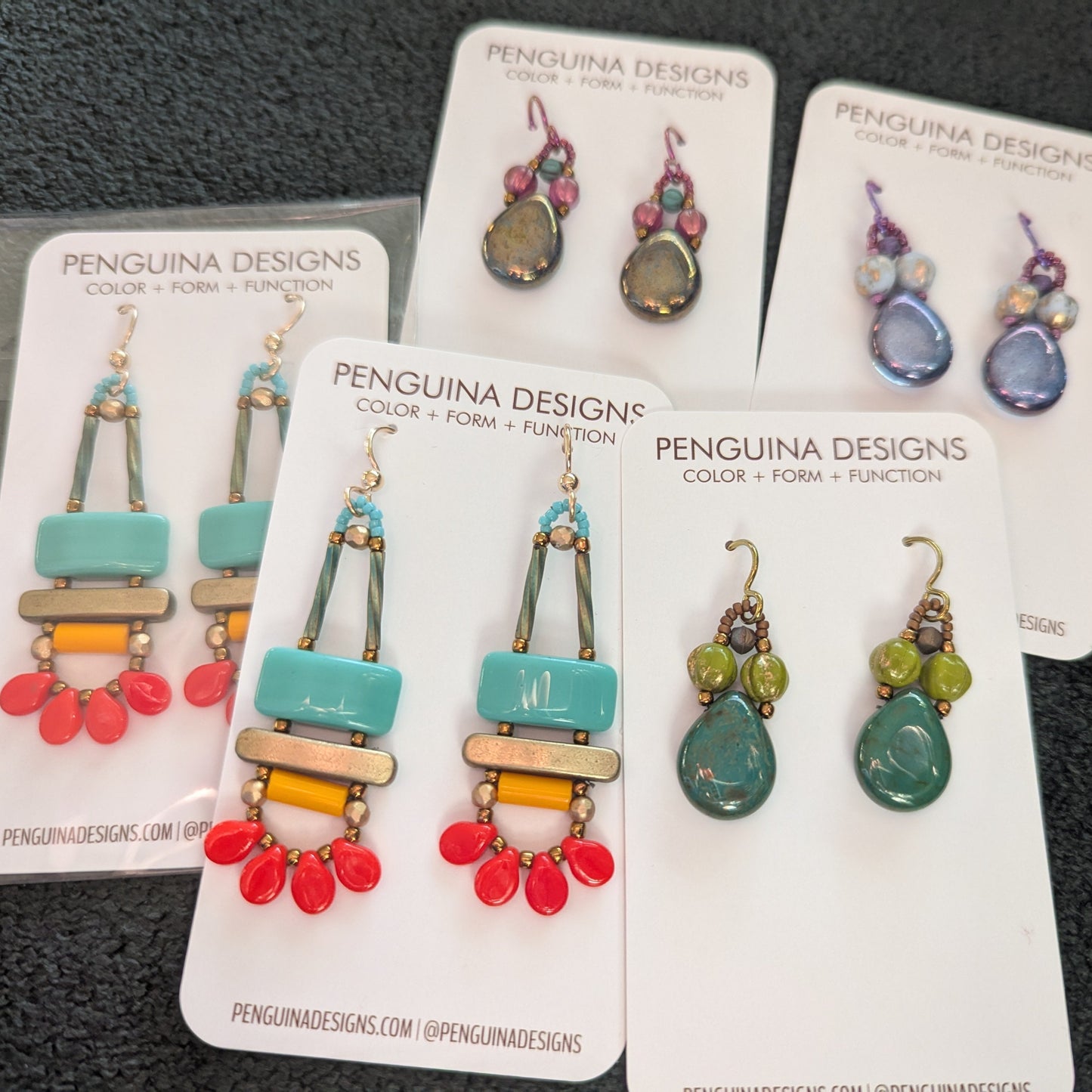 Five pairs of earrings on white cards are spread out on a fuzzy black background. Two pairs are bigger, with bright turquoise rectangles that have a gold bar below them and a swag of red petal beads. The other three are all one style, big teardrops topped by a pair of round beads and one smaller at the top. One pair is pink and lustered green, one is clear blue with pale opaque blue and pink, and one is dark forest green with lime and gold.