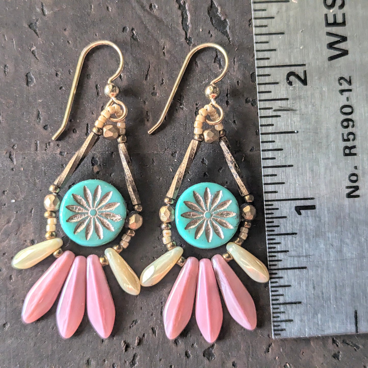 Pair of earrings with turquoise center and pink fringe on a dark surface with a ruler for scale.