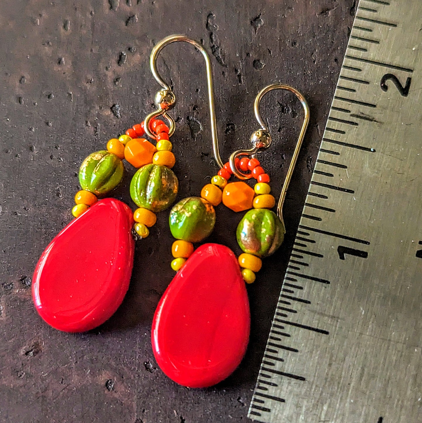 Colorful beaded earrings with red teardrop-shaped beads accented with green, yellow and orange beads on a textured surface with a ruler for scale.