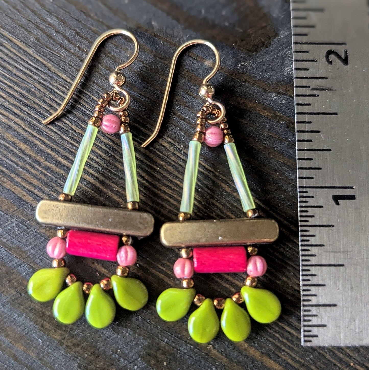 Colorful earrings with green, pink, and gold elements on a dark surface with a ruler for scale.