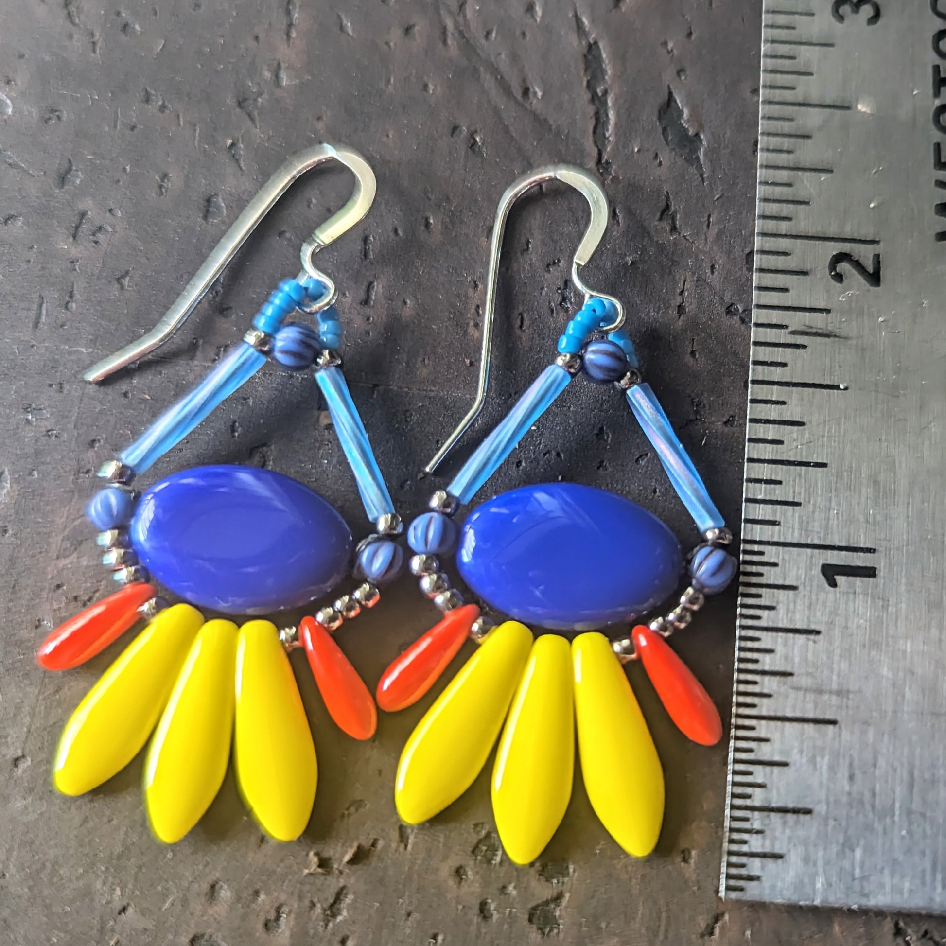 Colorful beaded earrings with blue, yellow, and red components on a dark surface with a ruler for scale.