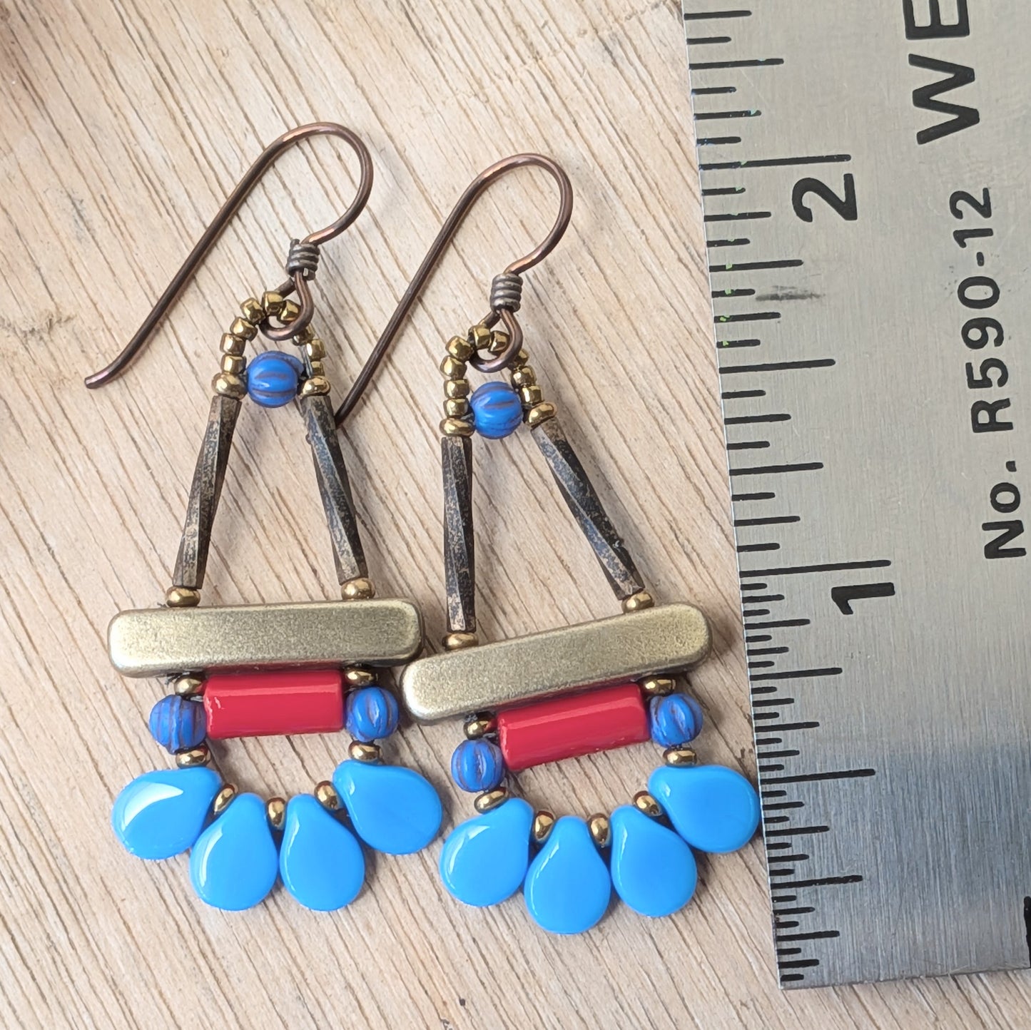 Blue and red earrings with petal shaped beads around the bottom, on a wood background with flowers and a ruler for scale.