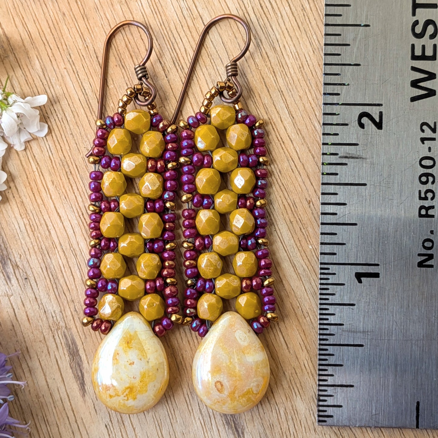 Yellow and purple beaded earrings with speckled cream teardrops at the bottom resting on a wood background with a ruler for scale.