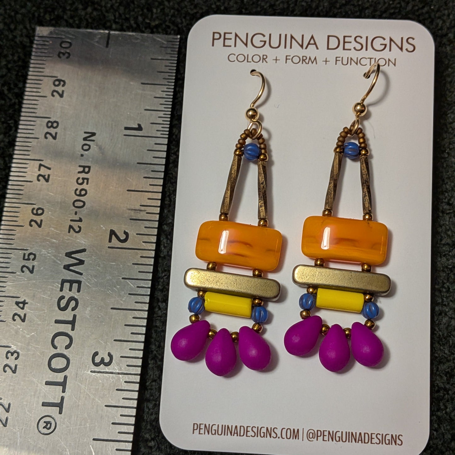 Colorful orange, yellow and fuchsia earrings a Penguinia Designs card with a ruler for scale.
