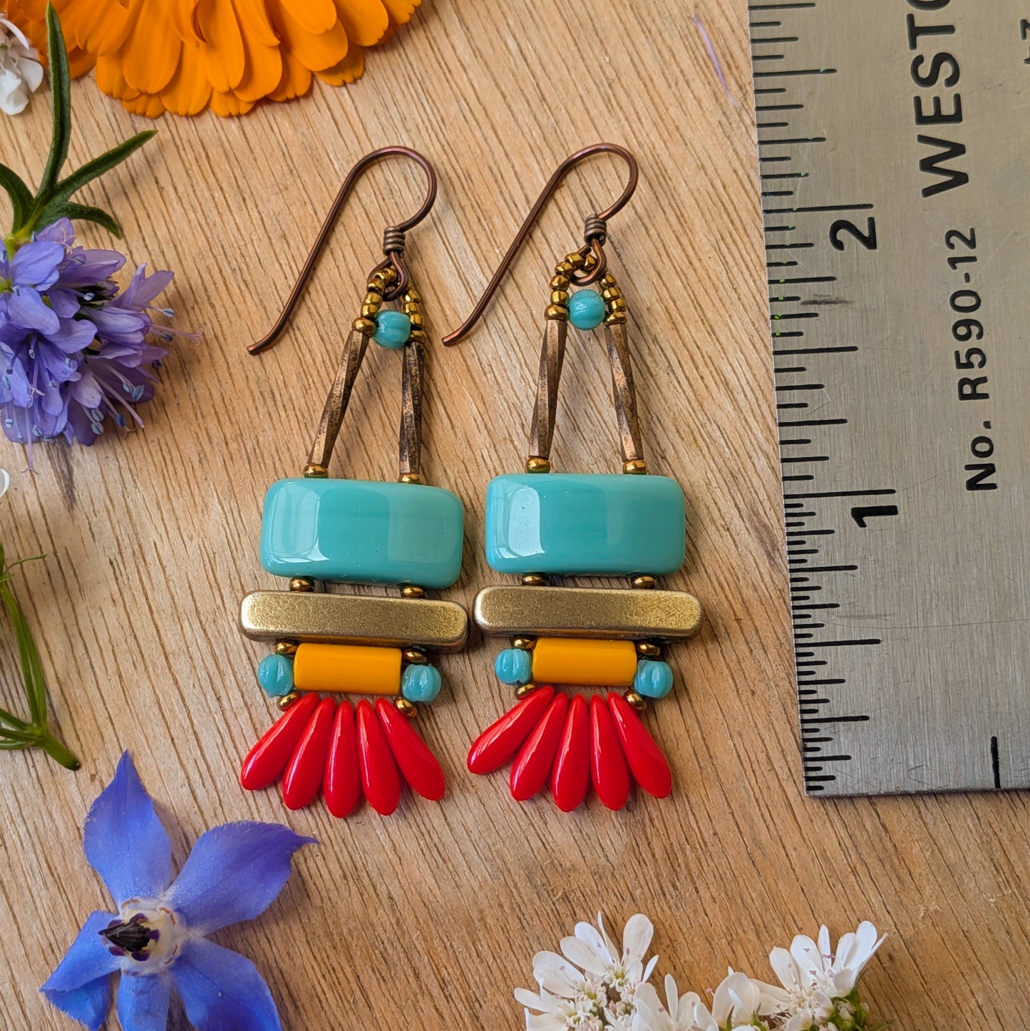 colorful earrings on a wood background with a ruler for scale. The earrings each have a wide turquoise rectangle stacked on top of a brass colored bar. Under the bar there is light orange tube, five small red daggers and tiny turquoise accents.