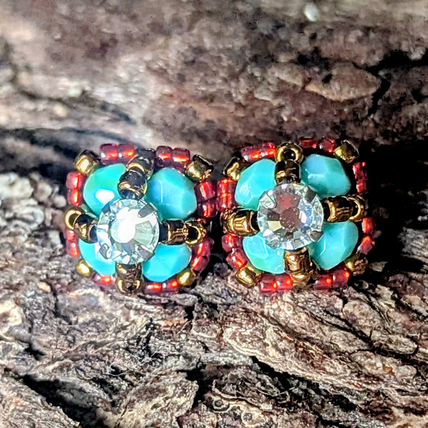Four-lobed stud earrings made with larger turquoise and smaller red beads and a rhinestone center, on a wood bark background.