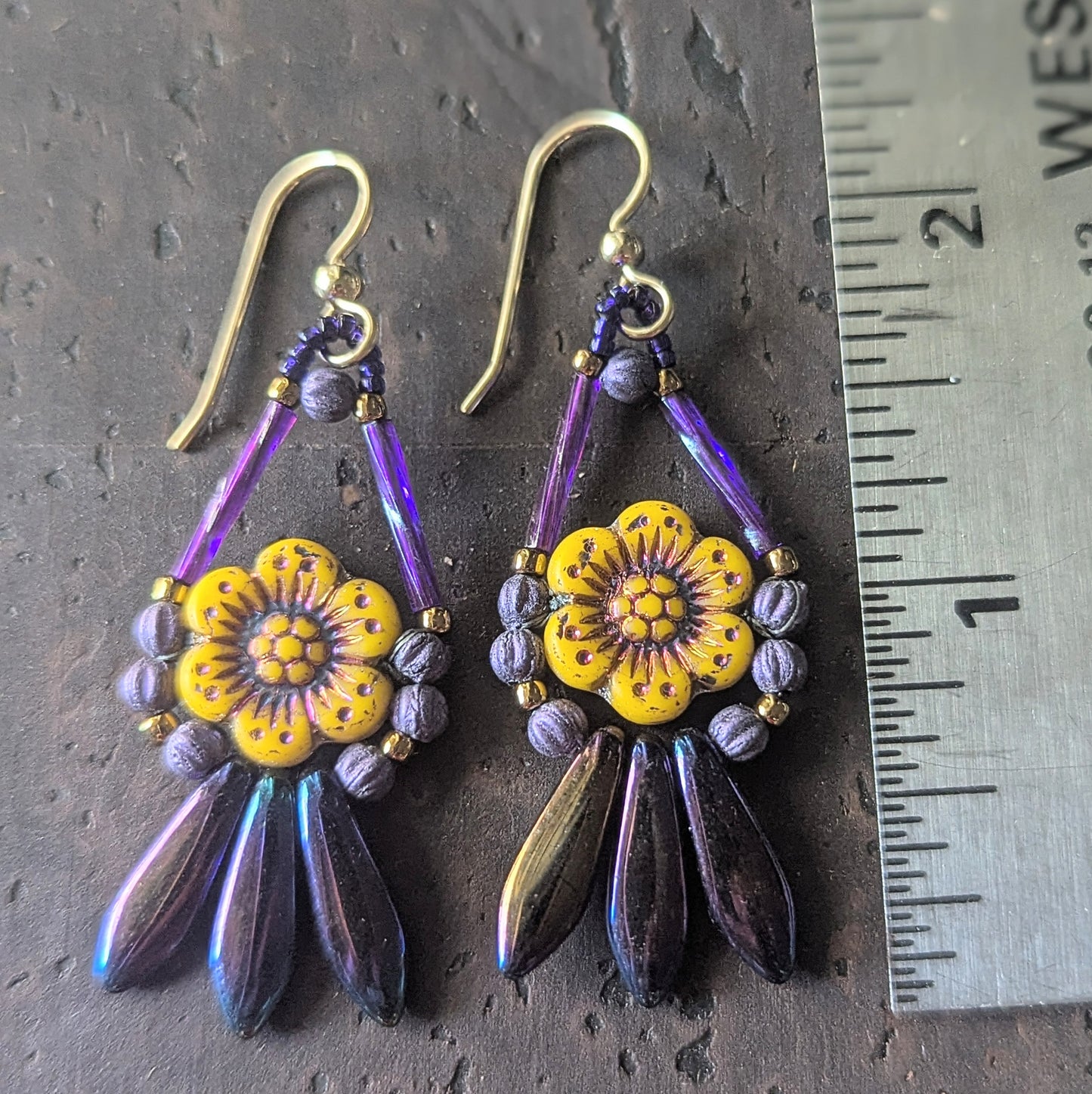 A pair of earrings with yellow flowers and a fringe of iridescent purple beads on a dark brown surface, shown with a ruler for scale.