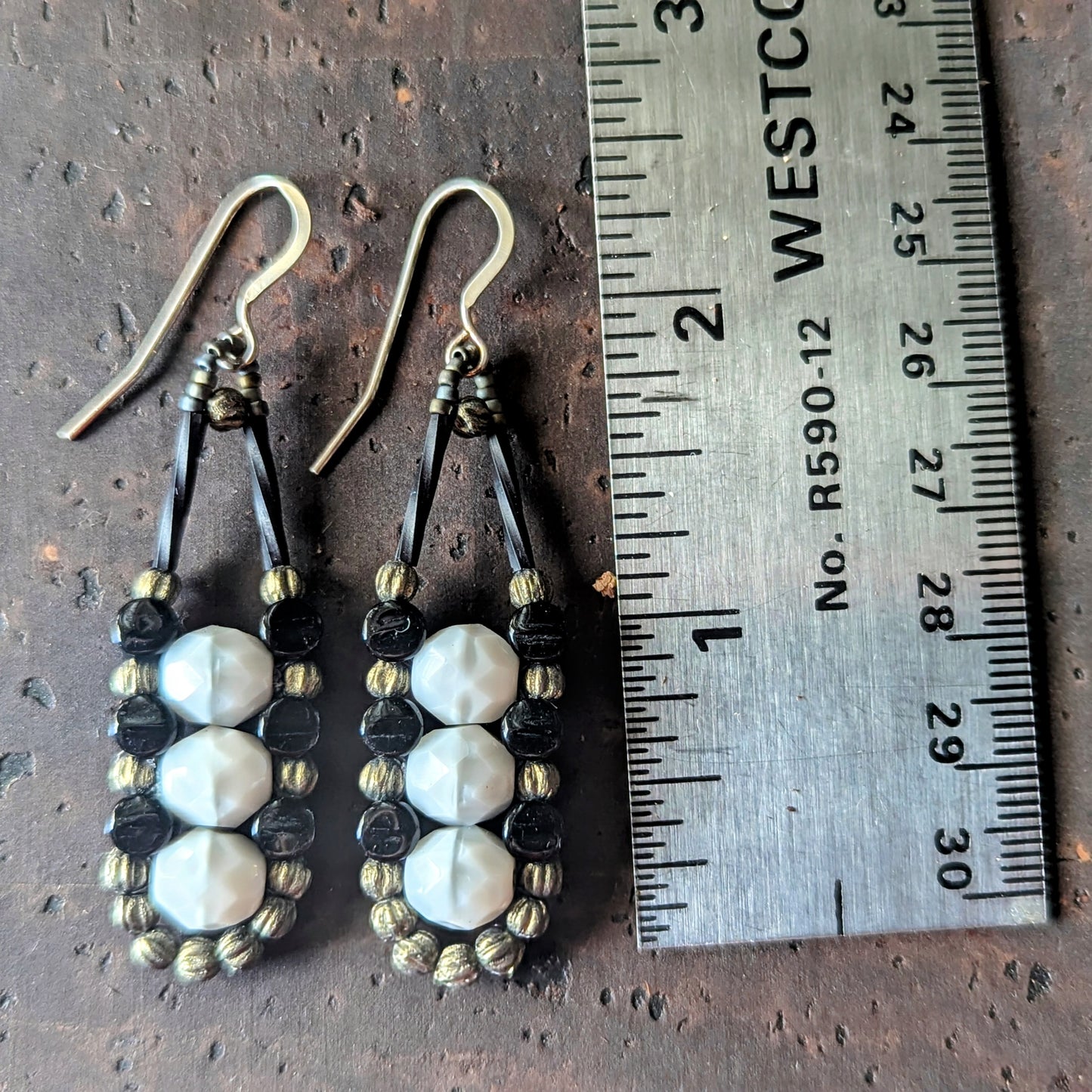 A columnar pair of gray and black beaded earrings next to a ruler on a textured brown surface.