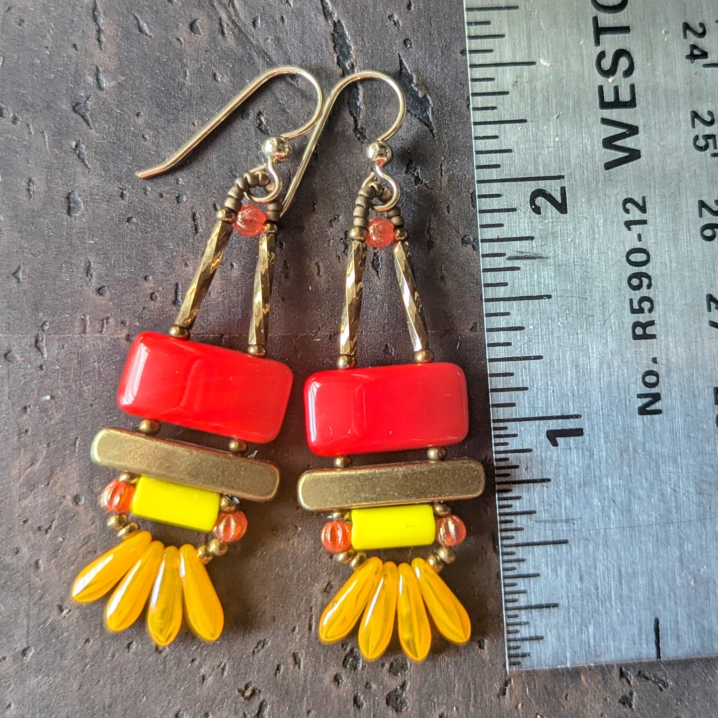 Colorful earrings with red, yellow, and gold elements on a dark surface with a ruler for scale.