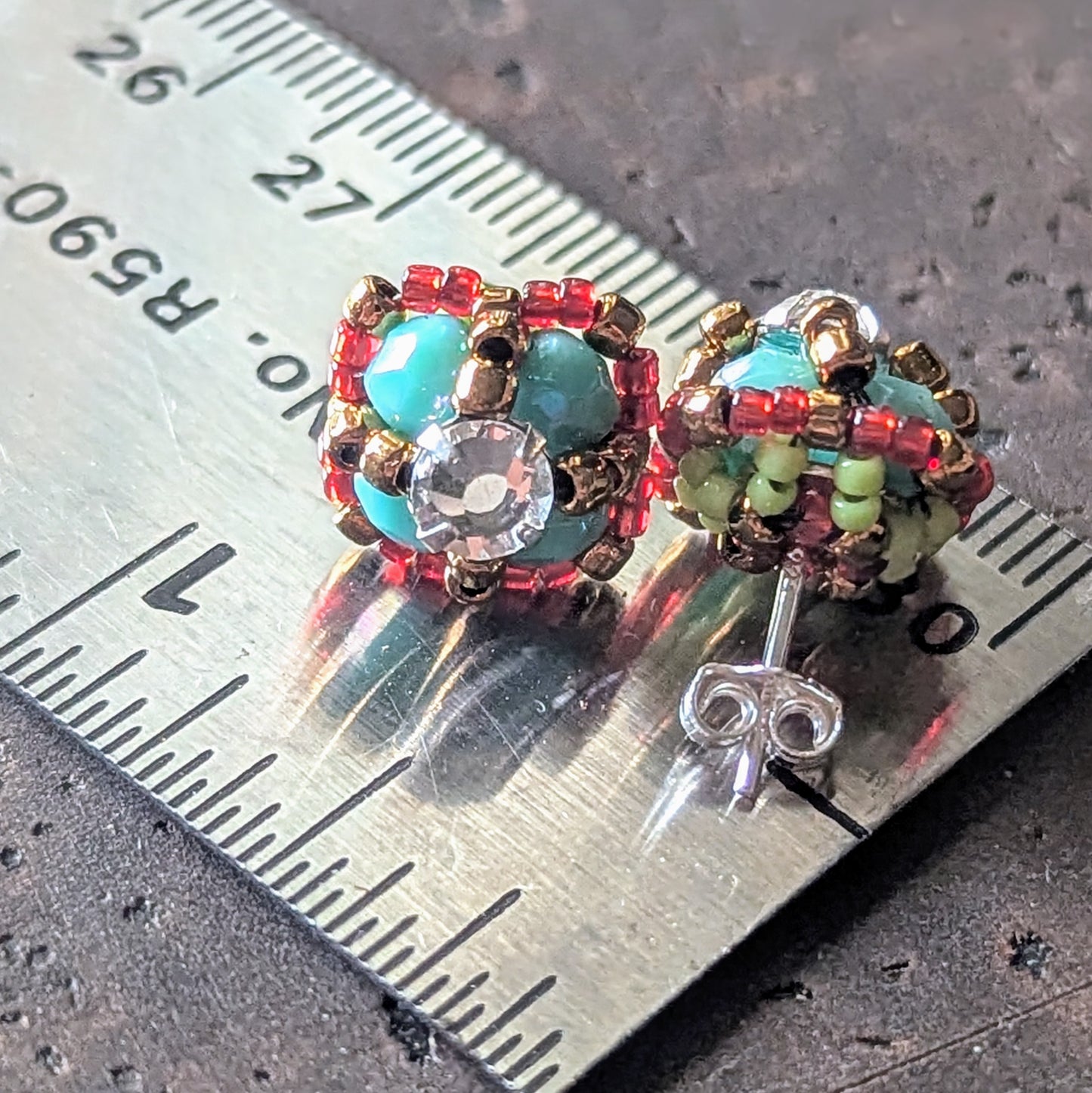 Four-lobed stud earrings on a ruler for scale. The earrings are made with larger turquoise beads surrounded by smaller red beads and a rhinestone center, shown with the back of one stud that has green details.