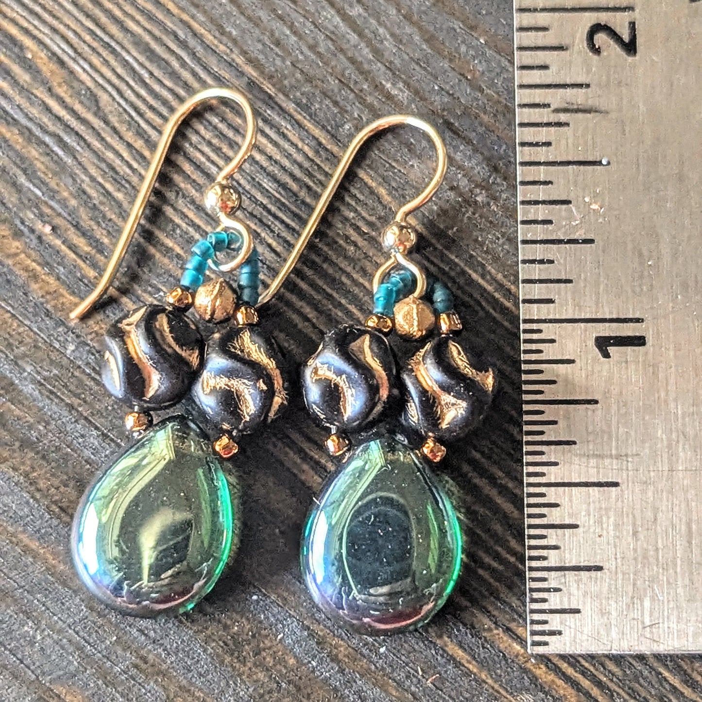 Pair of earrings with green teardrop-shaped beads and gold accents on a wooden surface with a ruler for scale.