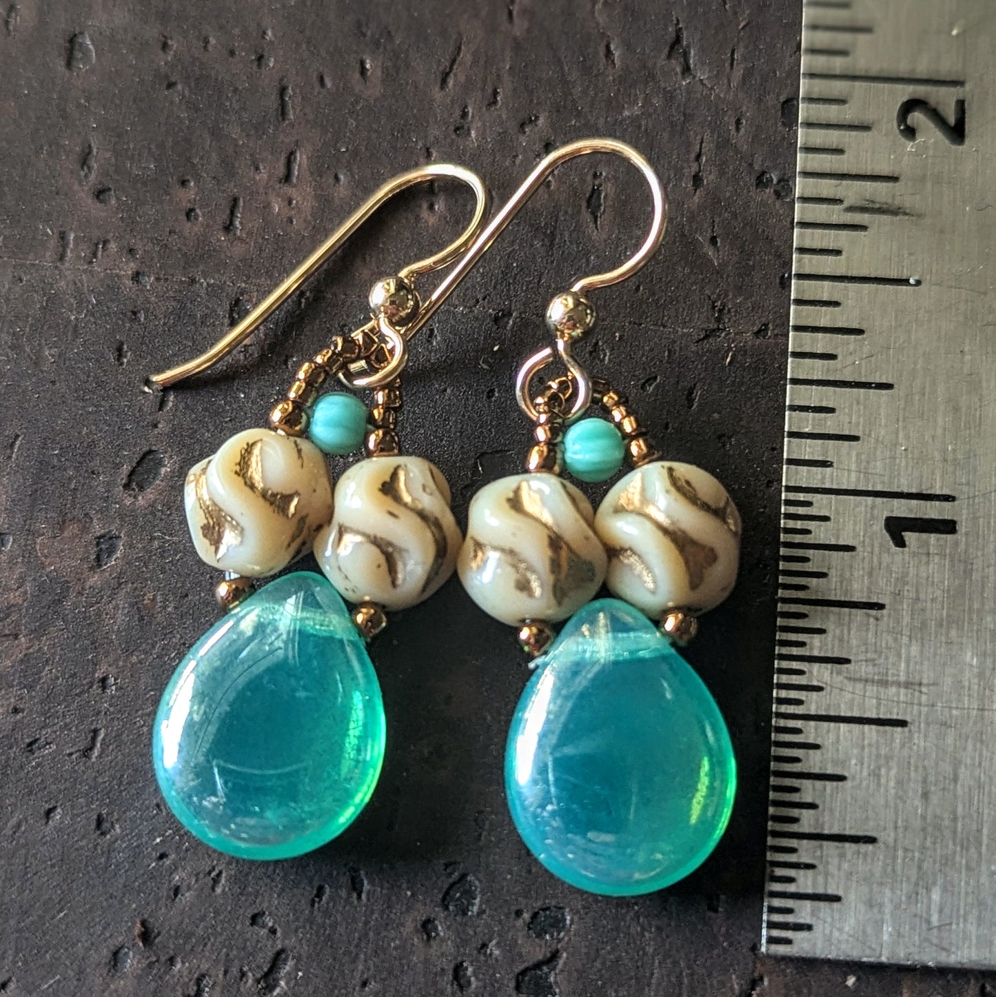 Pair of earrings with lustered blue-green teardrops on a dark surface with a ruler for scale.