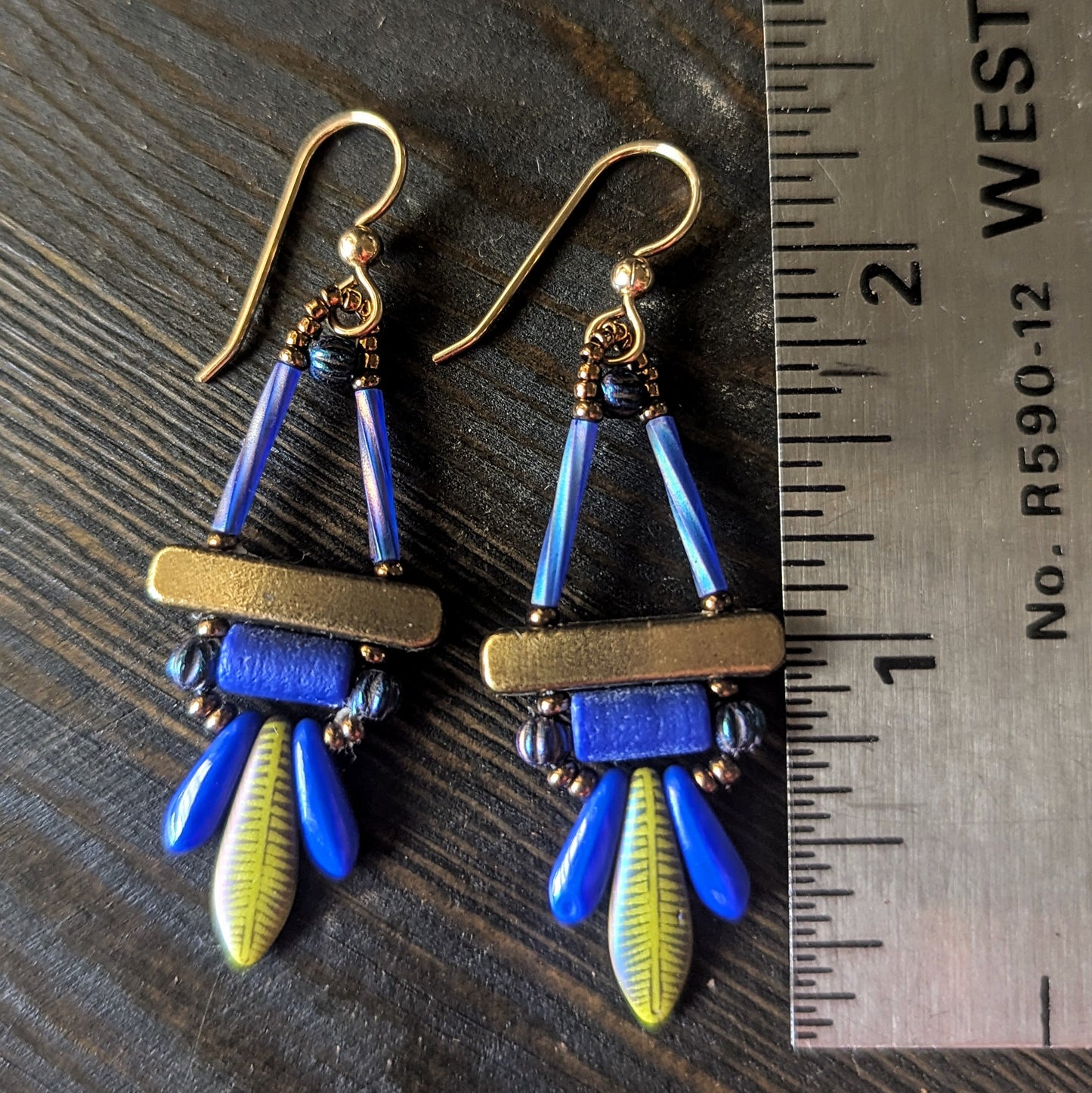 A pair of colorful earrings with gold, blue and olive green components next to a ruler on a wooden surface