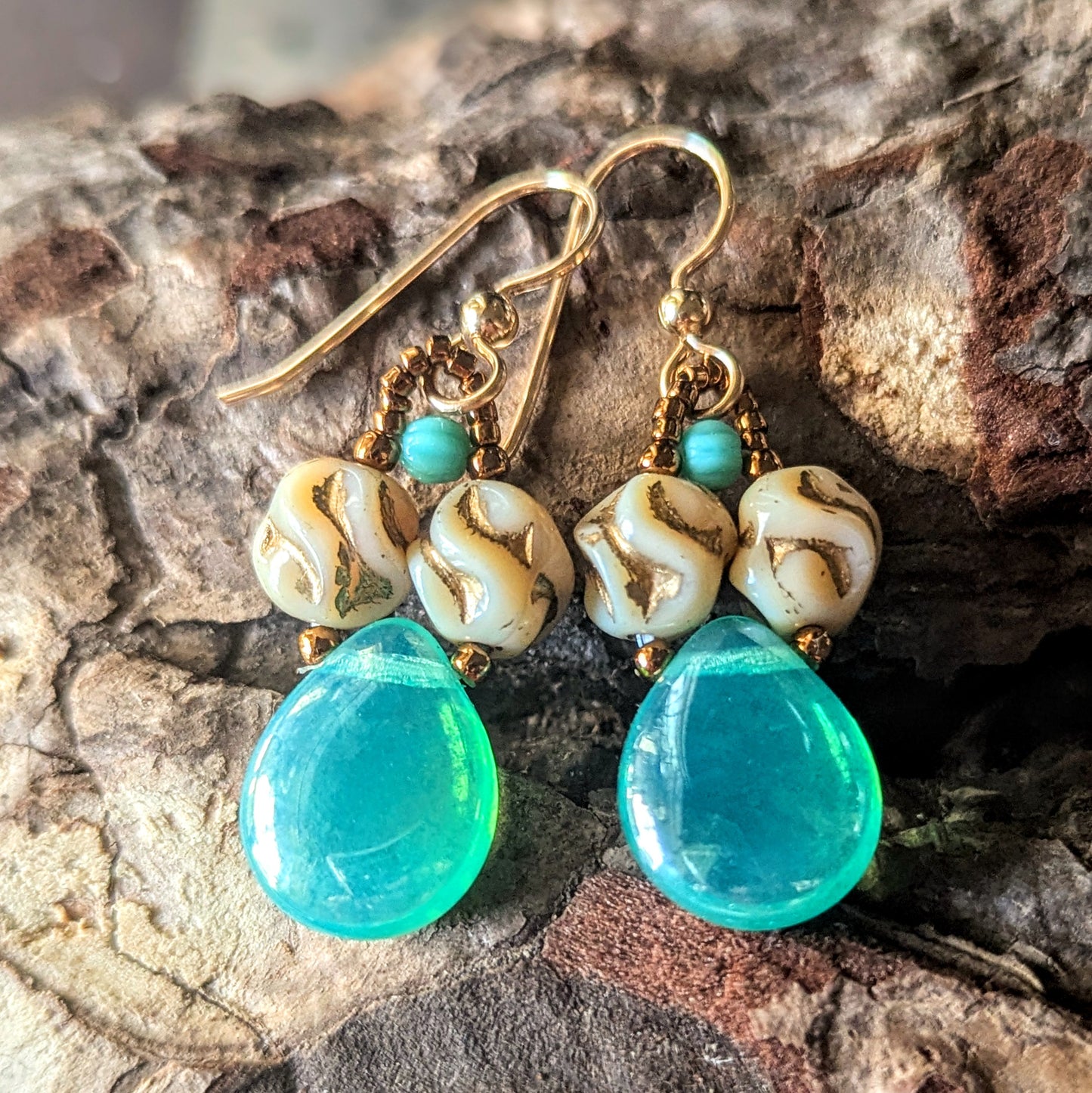 Pair of earrings with lustered blue-green teardrops and gold accents on a textured surface