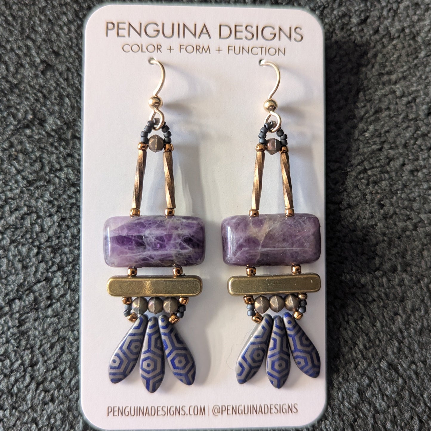 Purple earrings on a paper card. The earrings have purple stone rectangles on top with gold bars and navy blue daggers with a silver-gray octagon pattern.