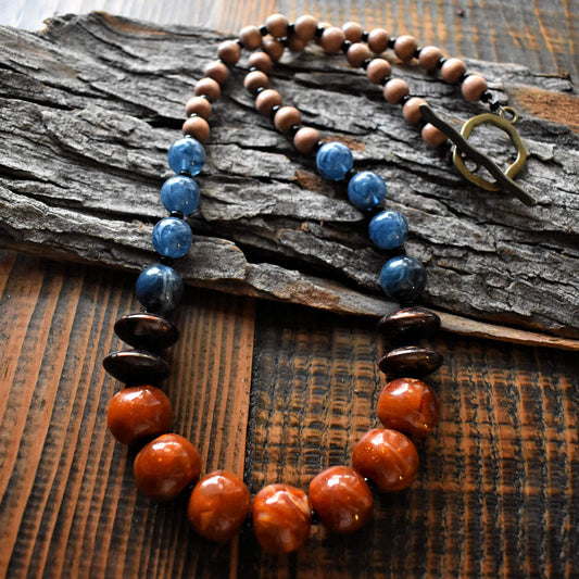 A blue and brown necklace with a large brass toggle clasp is draped across a wood background. The necklace has seven large rust colored beads with some swirls of glitter. A pair of swirled chocolate discs are on either side, followed by a series of four swirled, transparent denim blue beads. The back half of the necklace is medium brown wood beads.