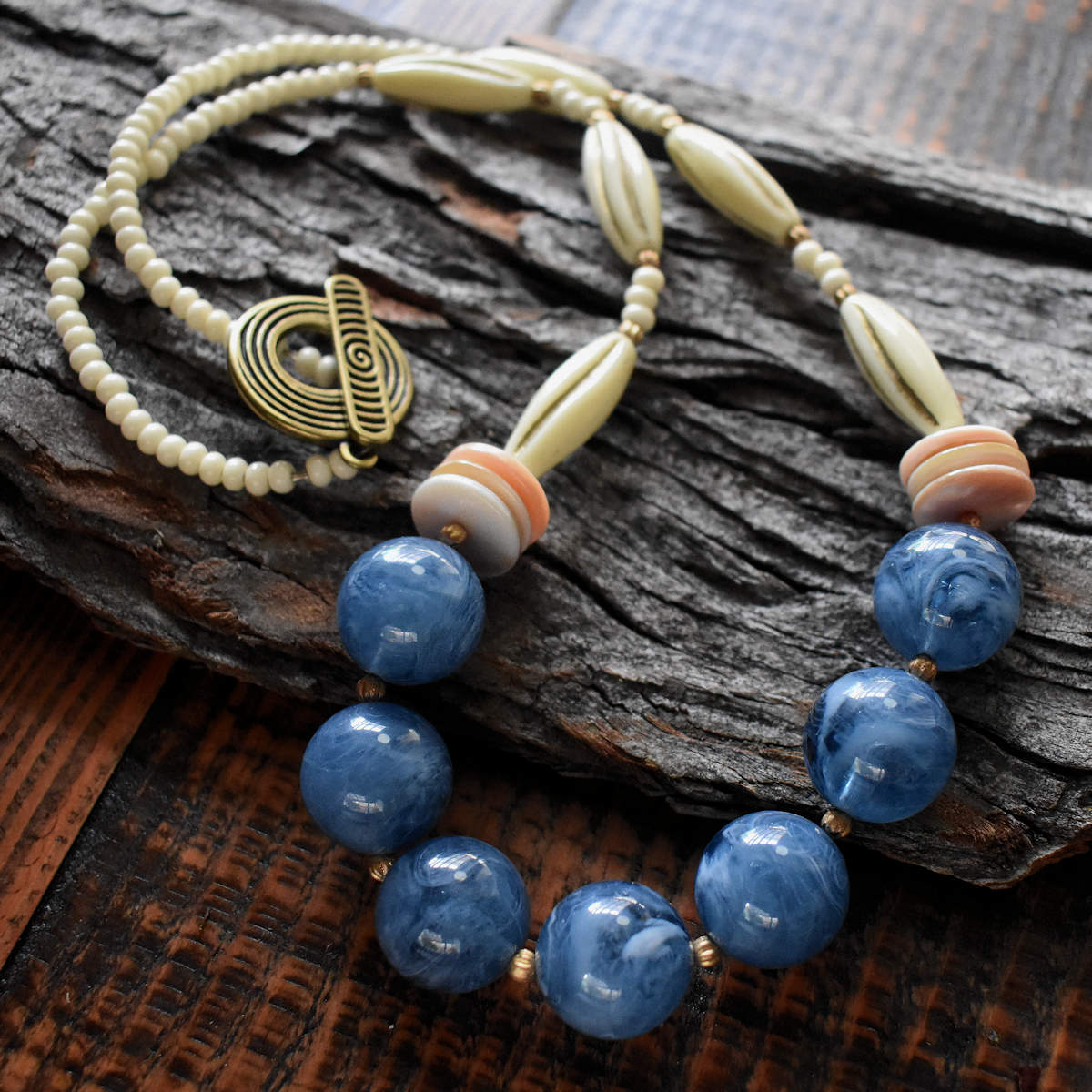 A cream and blue necklace is draped in a squiggle over a wood background. The necklace has chunky swirled blue beads at the center, a stack of three peachy pink shell discs, and then three long cream beads with gold embossed lines. The back of the necklace is chunky cream seed beads and a gold colored toggle clasp with a dramatic spiral pattern enhanced with black.