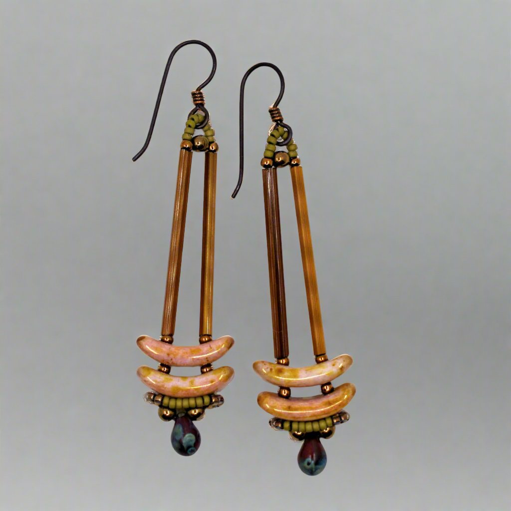 Earrings that look similar to a medicine dropper lay against a gray background. There are two long, brown beads forming narrow vertical columns ending at the top of two speckled pink stacked arc shaped beads. At the bottom is a speckled red drop shaped bead, surrounded by a belt of gold seed beads.