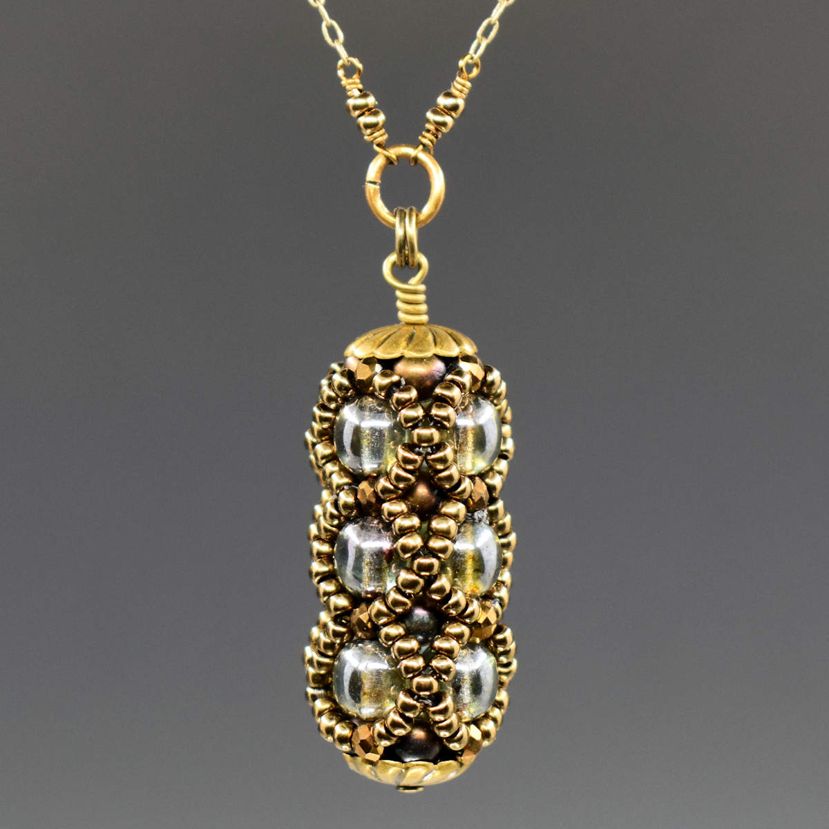 A small columnar pendant in gold and olive green hangs from a golden chain. The primary beads used are three stacked rows of lustered olive green rounds, surrounded by a netting of gold seed beads.