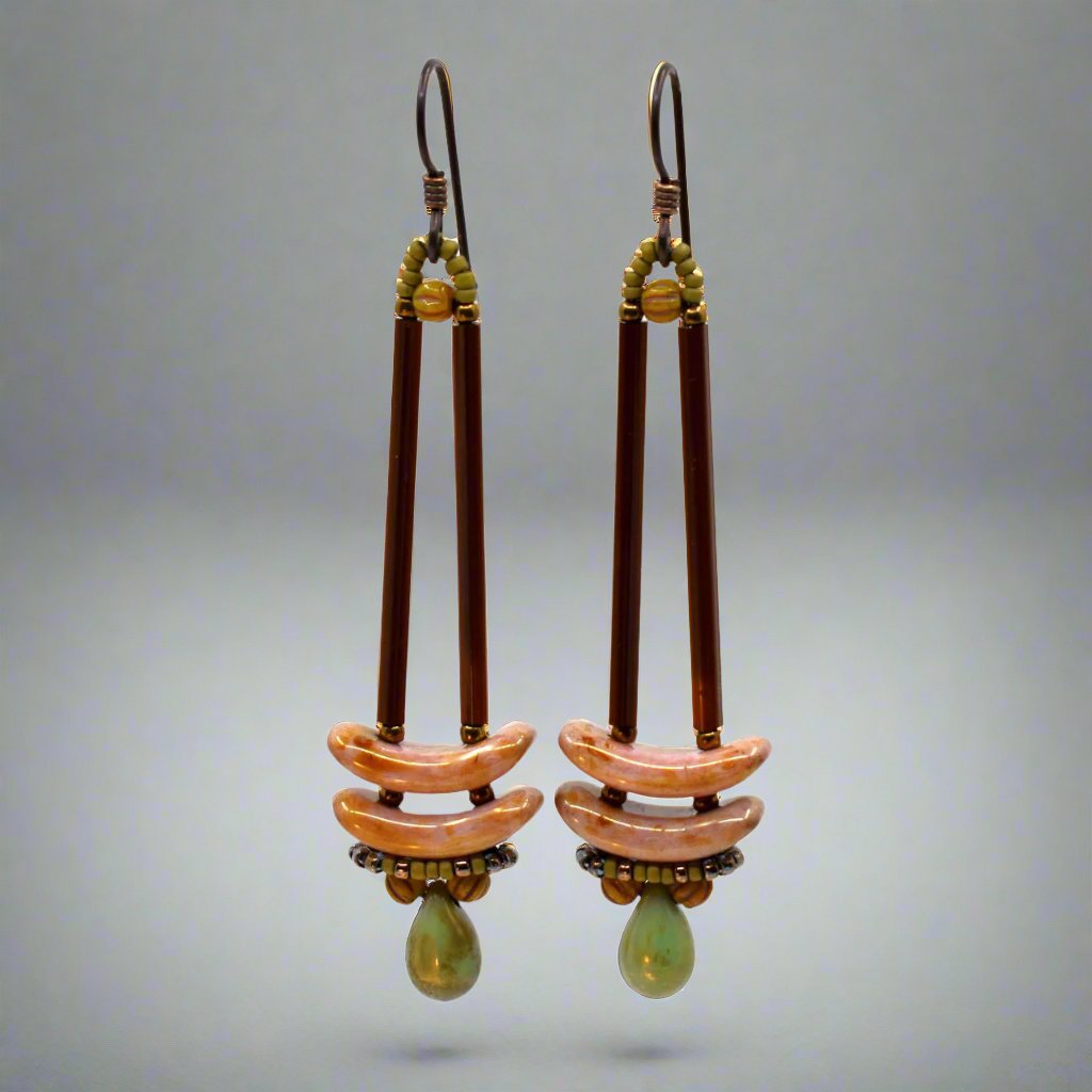 Earrings that look similar to a medicine dropper lay on a white backround. There are two long, brown beads forming narrow vertical columns ending at the top of two speckled pink stacked arc shaped beads. At the bottom is a matte speckled turquoise green drop shaped bead, surrounded by a belt of matte gold seed beads.