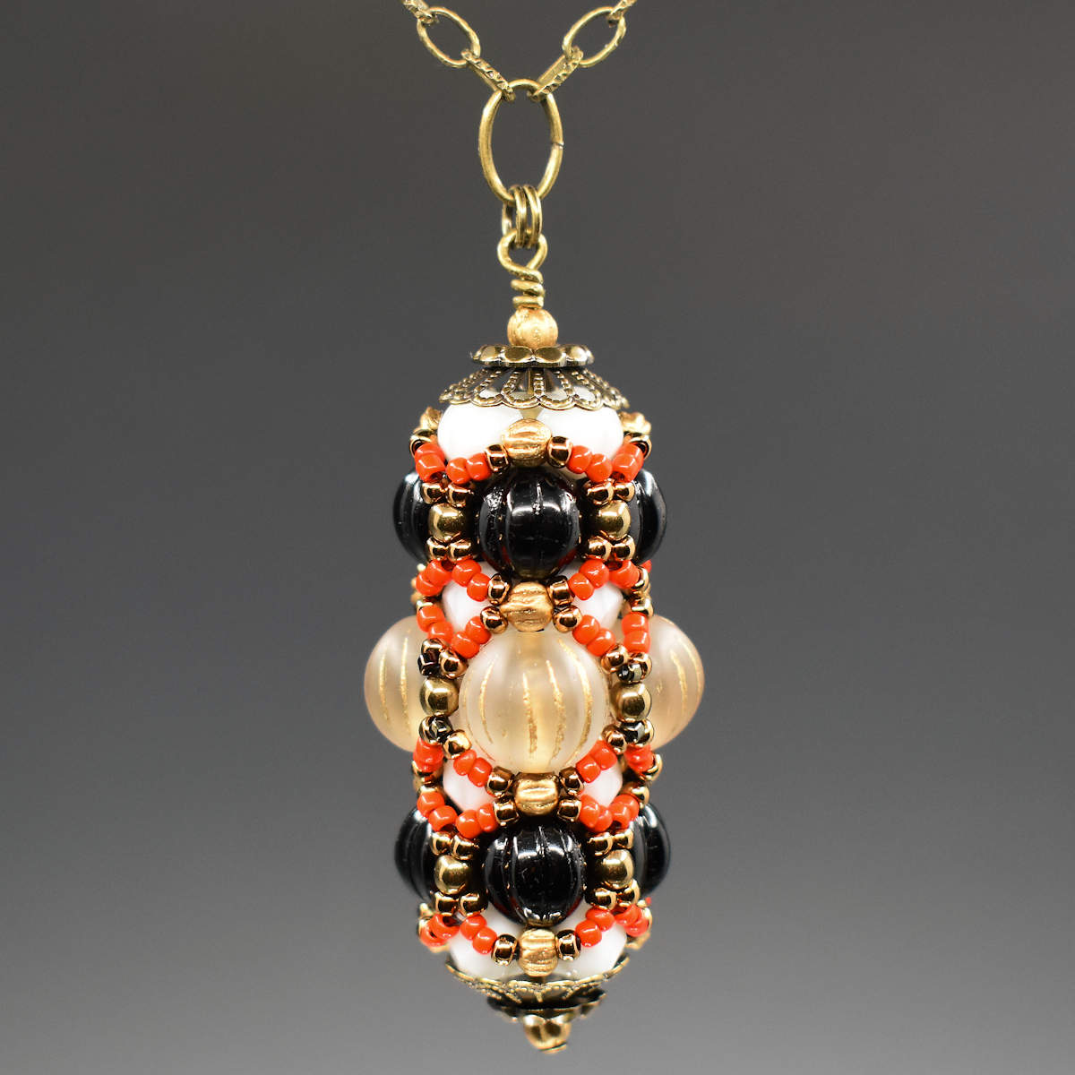 A black and cream pendant with coral accents hangs on a golden chain against a gray background. The pendant has a wider center row with frosted clear beads that have vertical gold stripes. The outer rows are both ridged black rounds. Smaller white beads helping form the structure are mostly hidden beneath the coral and gold seed bead netting that overlays the whole pendant.